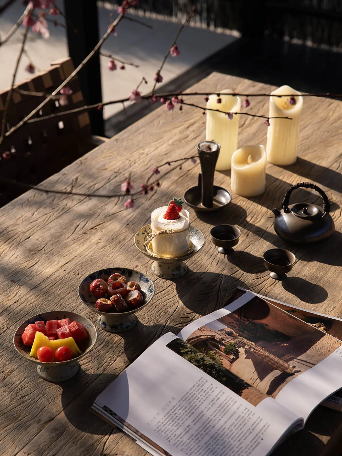 A rustic reclaimed wood table set with a small strawberry cake, fresh fruit, dried dates, a black cast iron teapot, ceramic teacups, white pillar candles, and flowering plum branches in warm sunlight.