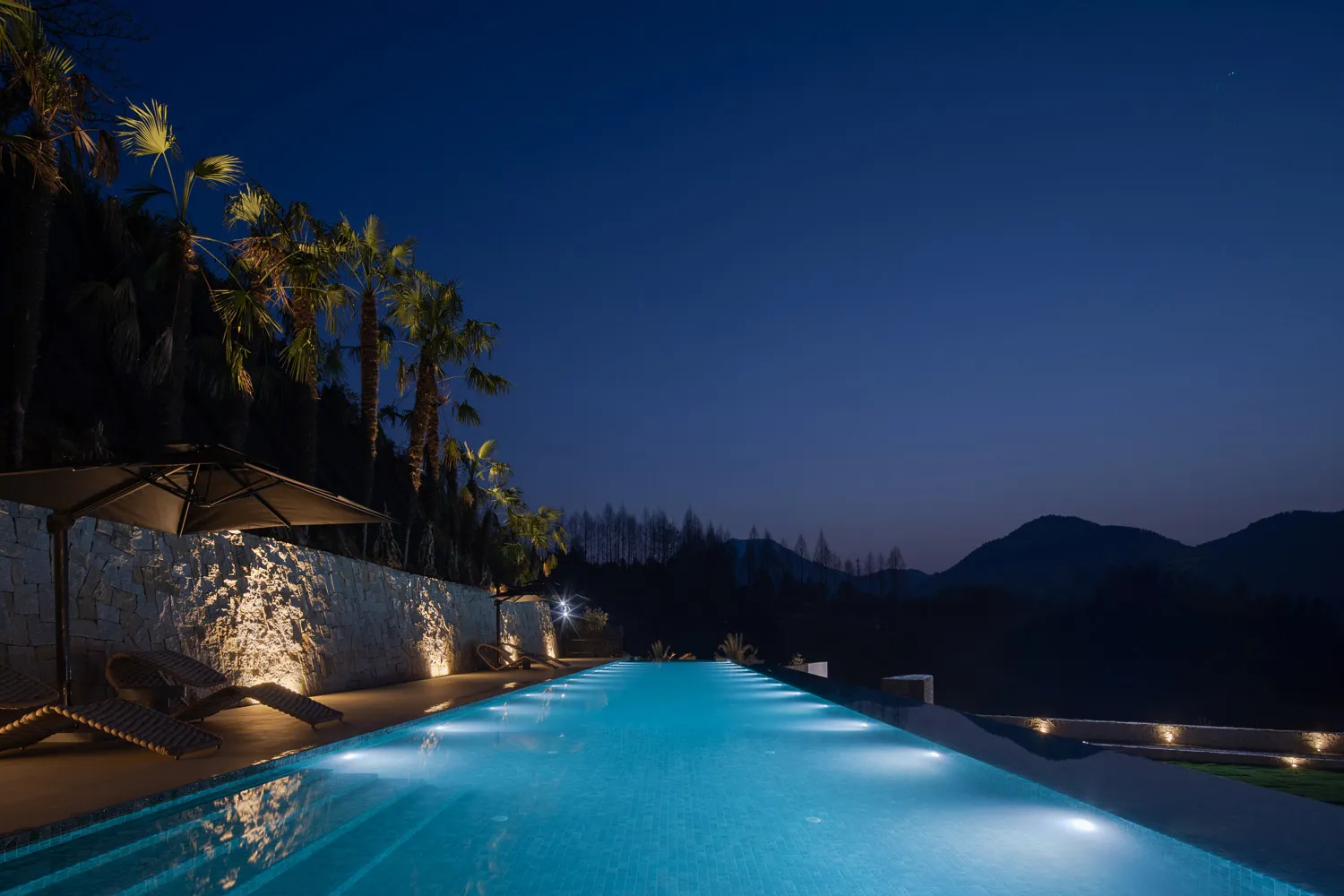 A luxurious infinity pool glowing with underwater lights at night, bordered by a textured natural stone wall, palm trees, and lounge chairs overlooking dark mountains.
