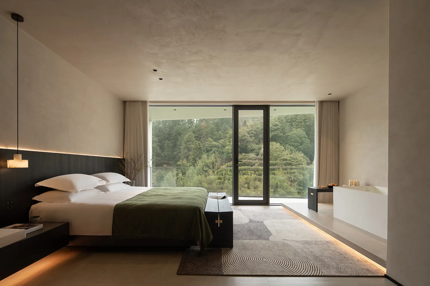 Modern minimalist bedroom featuring dark wood headboard, white bedding with green throw, concrete walls and ceiling, large floor-to-ceiling windows overlooking a dense forest, and a freestanding white bathtub on a raised platform.