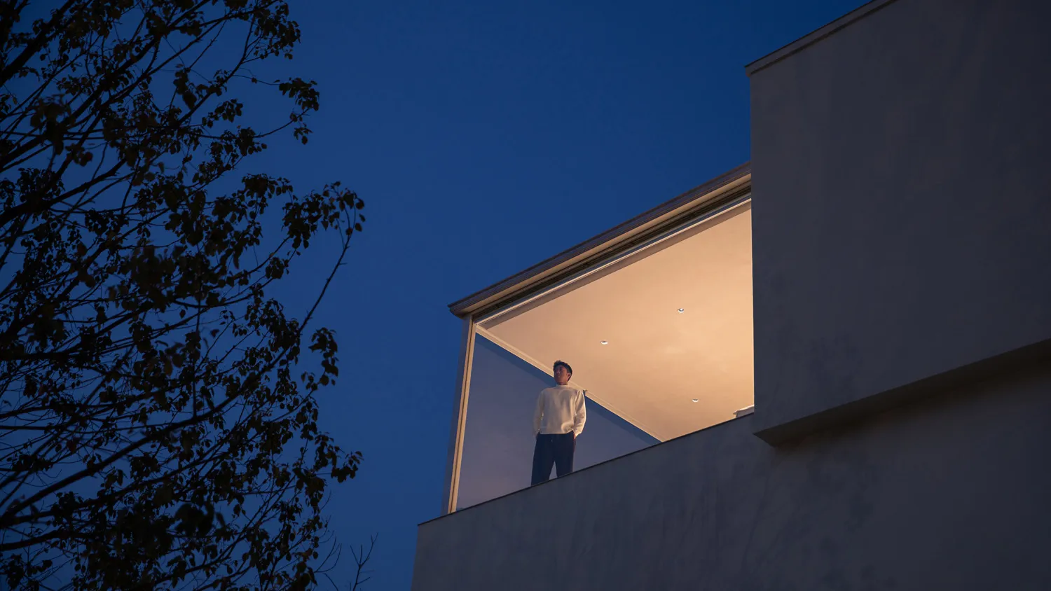 Man standing on a modern cantilevered concrete balcony at dusk with warm interior lighting against a deep blue sky and silhouetted tree branches.