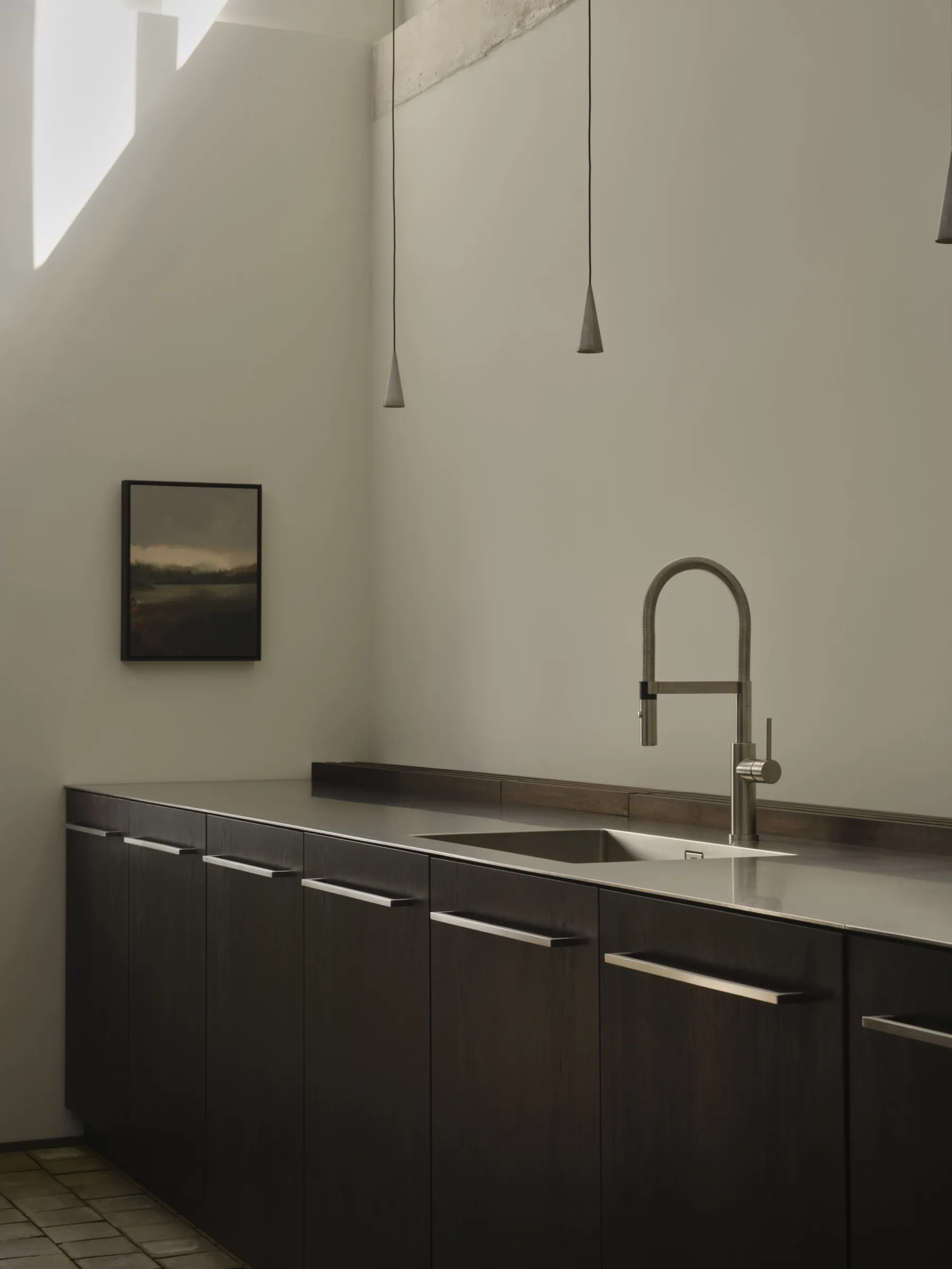 Modern minimalist kitchen featuring dark wood lower cabinets, stainless steel countertop and sink, high-arc faucet, concrete beam detail, and pendant lights with a framed landscape painting on the wall.