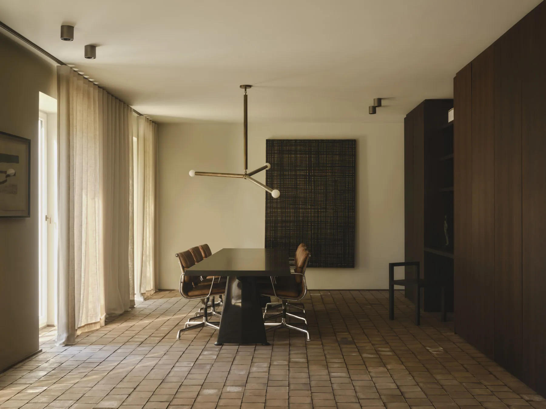 Modern minimalist dining room featuring a dark dining table, brown leather chairs, walnut wood paneling, large woven wall art, and light terrazzo tile flooring with sheer curtains allowing natural light.