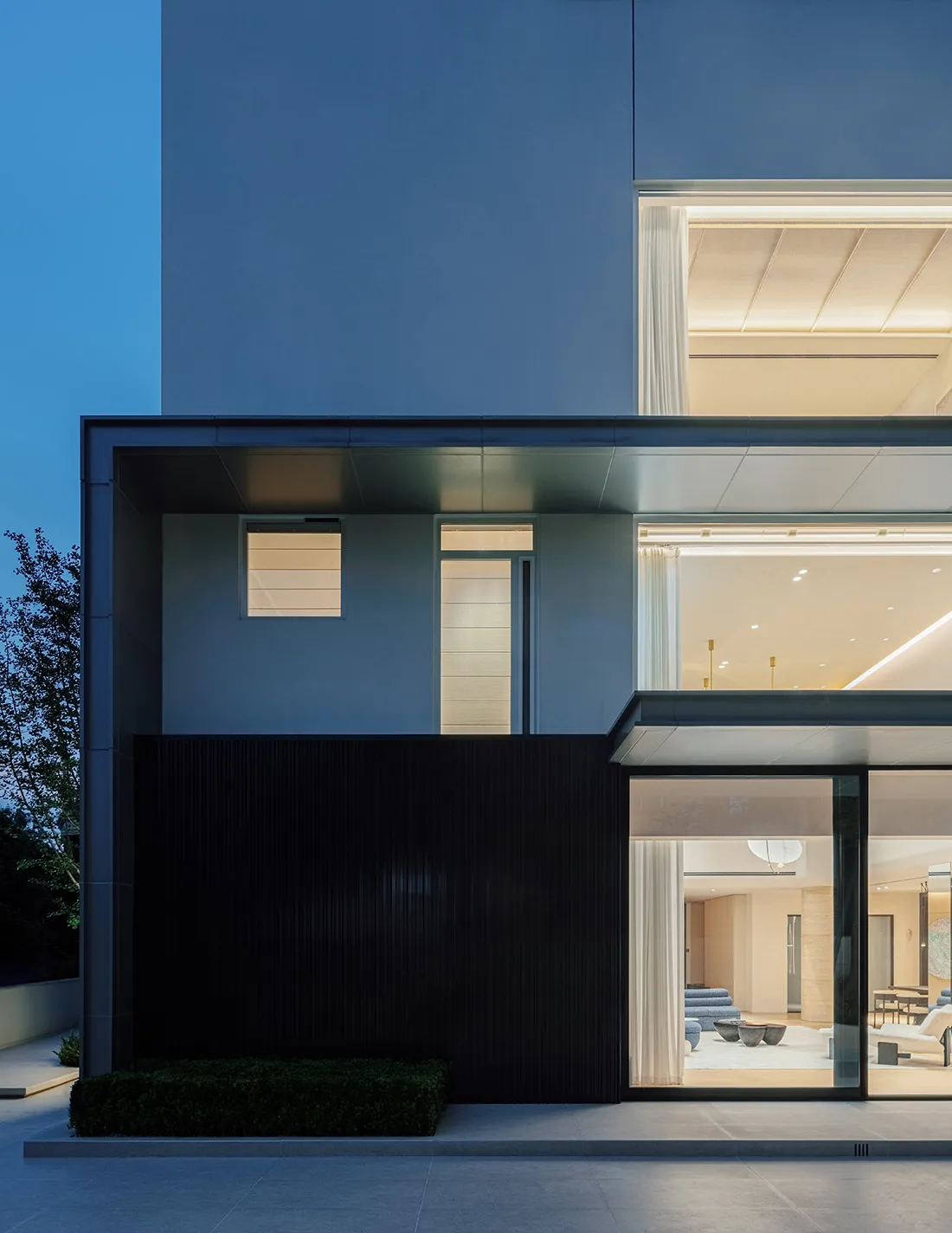 Exterior view of a contemporary luxury villa at dusk, showcasing dark wood paneling, large glass openings revealing an illuminated interior, and clean concrete architectural lines.