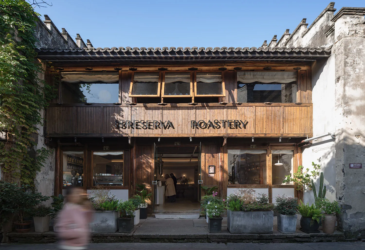 Street view of a two-story heritage building converted into 18 Reserva Roastery cafe, featuring dark reclaimed wood facade, large windows, open entrance, and numerous potted plants lining the stone pavement under a clear blue sky.