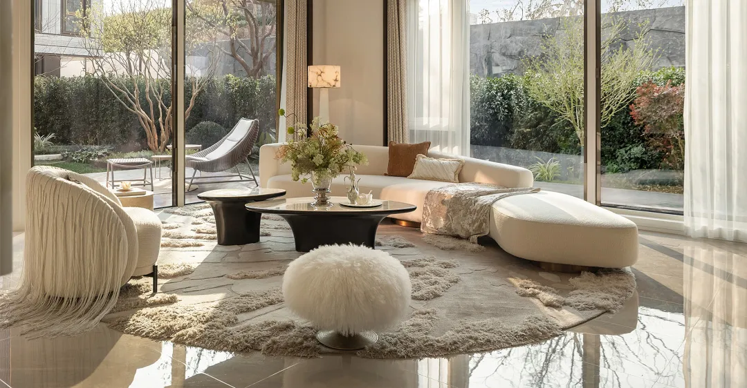 Bright modern living room featuring a curved white boucle sofa, cream fringe armchair, black coffee table, and white fur ottoman on a textured area rug with large glass doors opening to a garden.