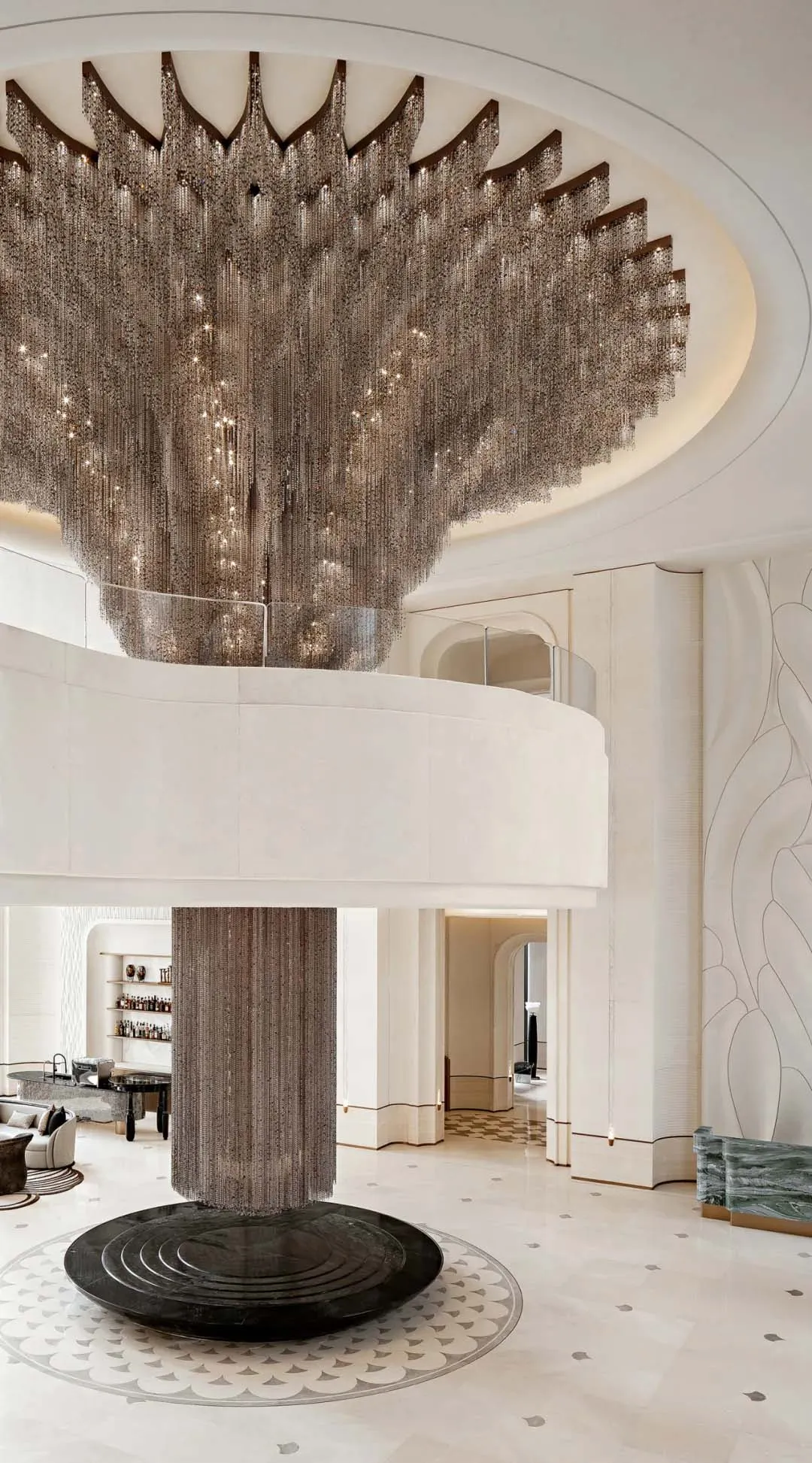 Expansive luxury hotel lobby featuring an enormous cascading crystal-bead chandelier hanging above a circular black marble base, with a curved mezzanine and light stone finishes.
