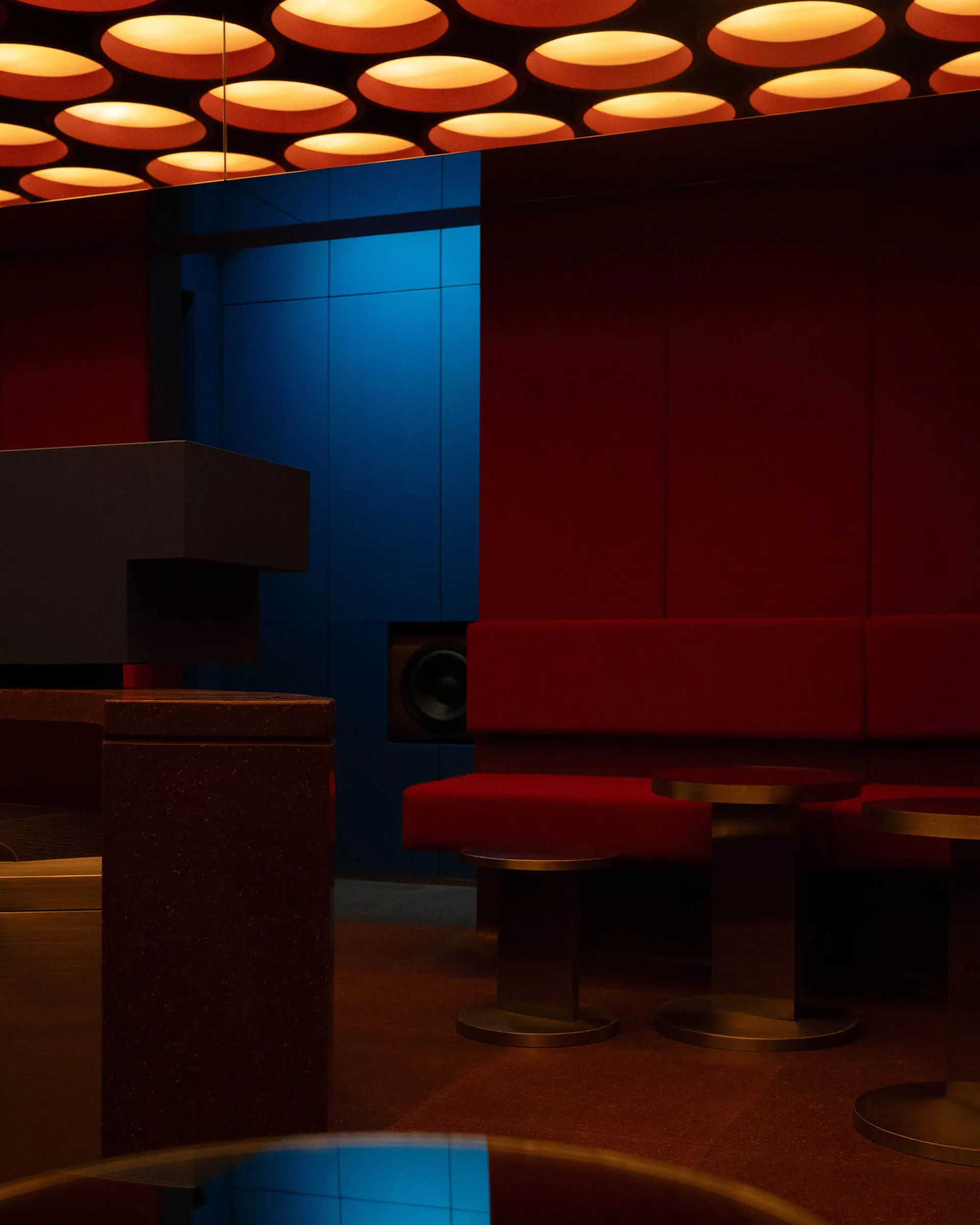 Interior view of a stylish nightclub lounge featuring red upholstered seating, dark terrazzo pedestals, circular warm ceiling lights, and vibrant blue backlit walls.