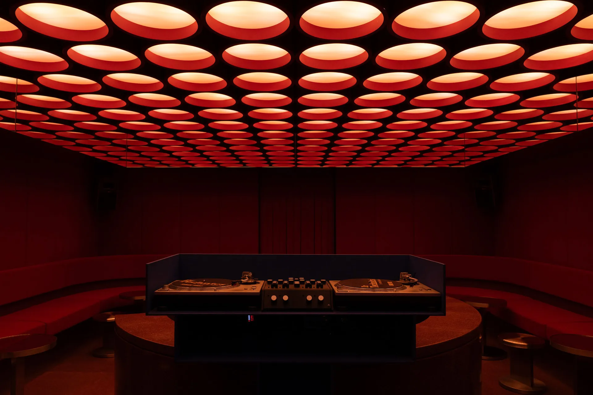 Interior view of a nightclub DJ booth featuring two turntables and mixer on a curved console, surrounded by red velvet curved banquettes under a ceiling covered with dozens of glowing red circular light panels.