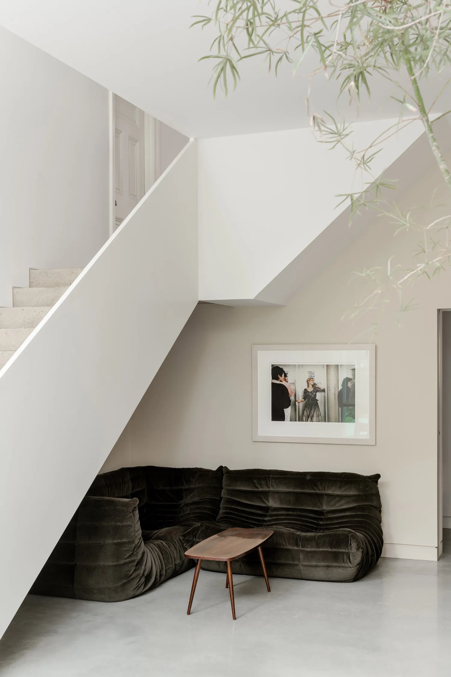Modern minimalist living space featuring a dark green corduroy modular sofa under a white staircase, accompanied by a walnut coffee table and framed black and white photograph on the wall.