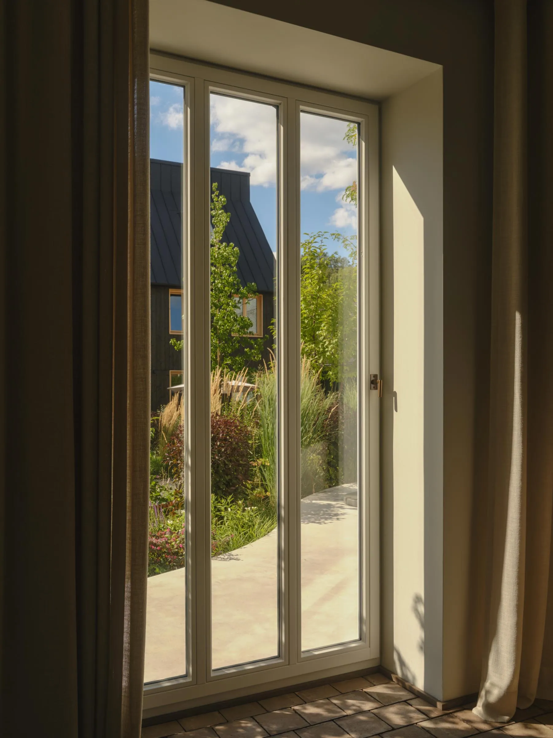View from inside a modern home looking out through a floor-to-ceiling triple glass door with light beige linen curtains on both sides, revealing a sunny garden path surrounded by tall grasses, flowering plants and a dark modern house in the background.