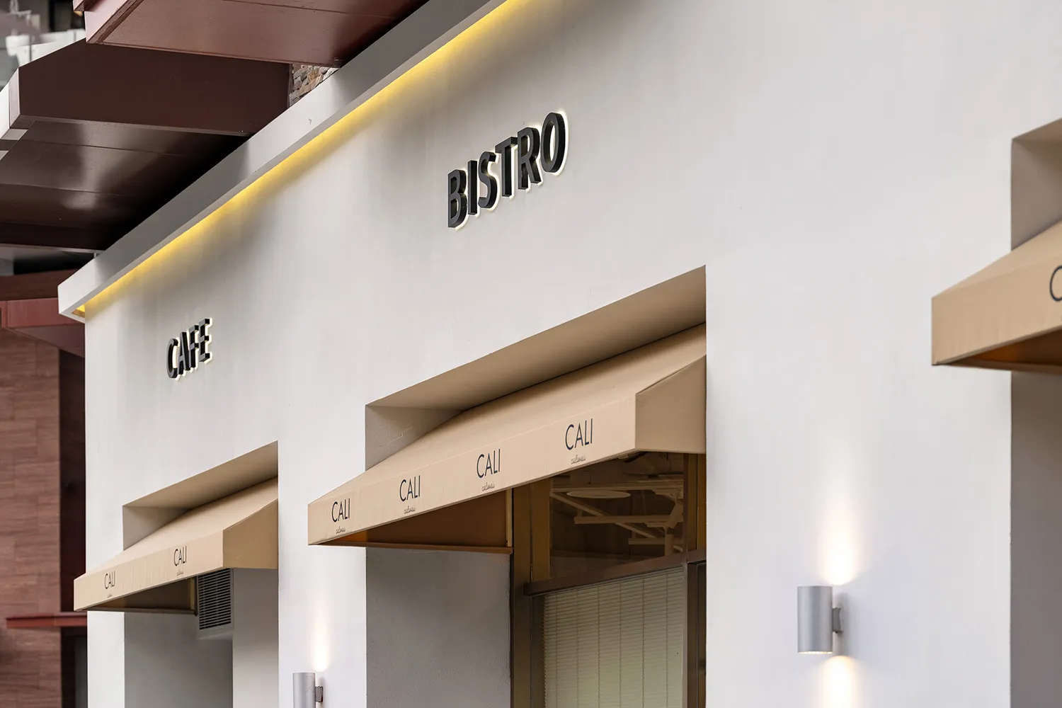 Modern white commercial facade of a cafe and bistro with beige fabric awnings, illuminated 'CAFE' and 'BISTRO' signs, and cylindrical wall sconces.