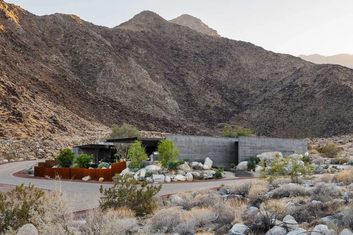Modern concrete house with large glass windows nestled among desert rocks and native plants, with a massive arid mountain range in the background at golden hour.