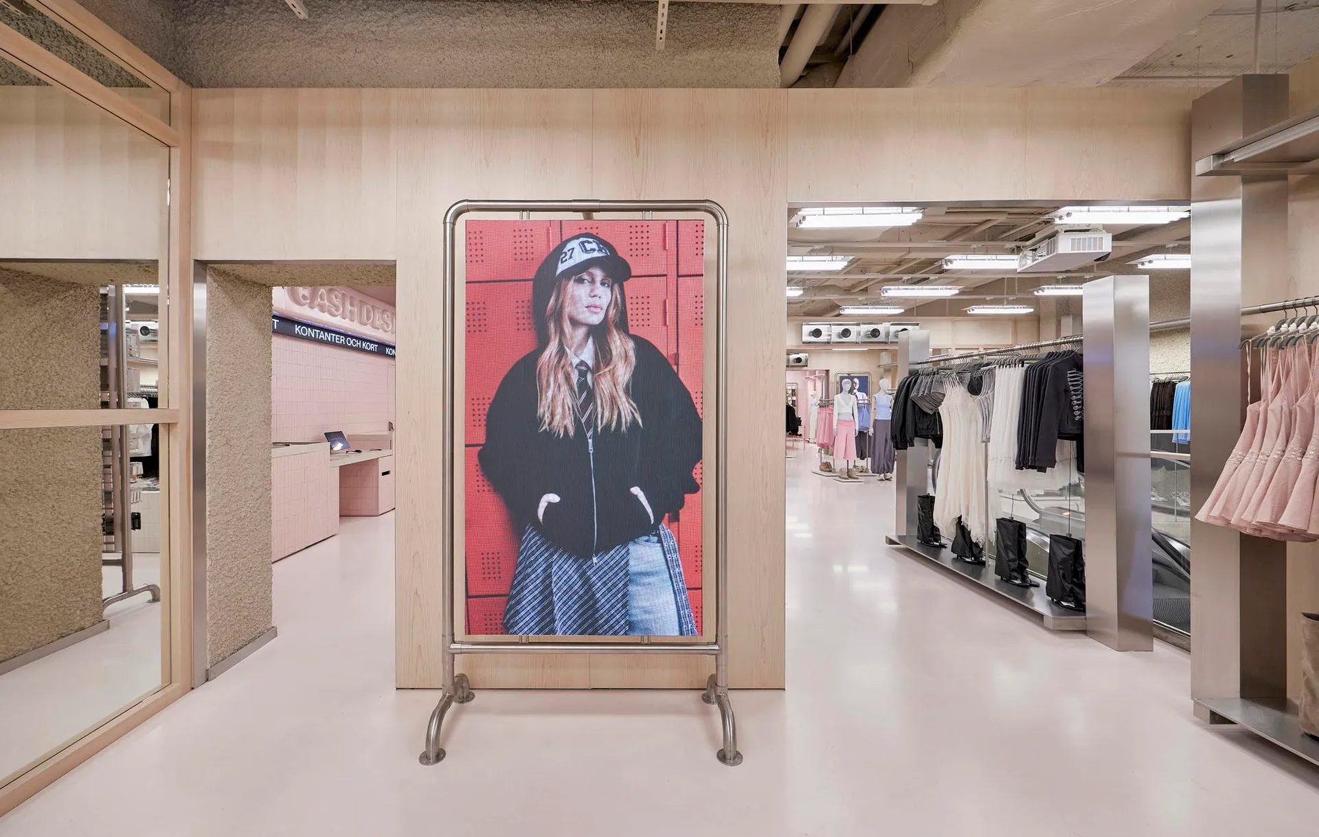 Modern fashion boutique interior featuring light wood paneling, a large digital portrait on a metal stand, stainless steel accents, and clothing racks in the background.