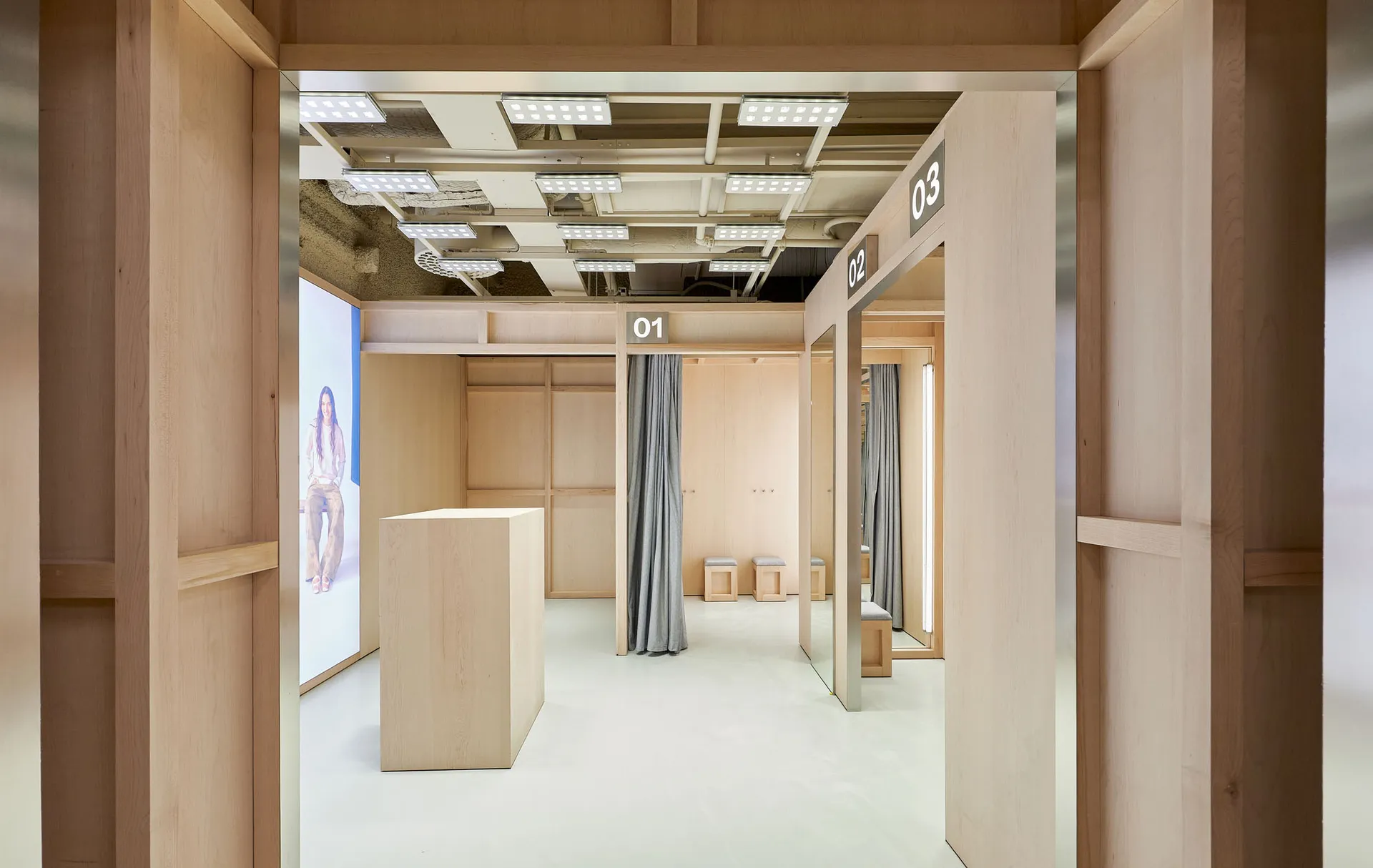 View into a modern minimalist retail fitting room area constructed with light-colored wood panels, featuring numbered changing rooms, grey curtains, a central wooden display block, and integrated LED lighting.