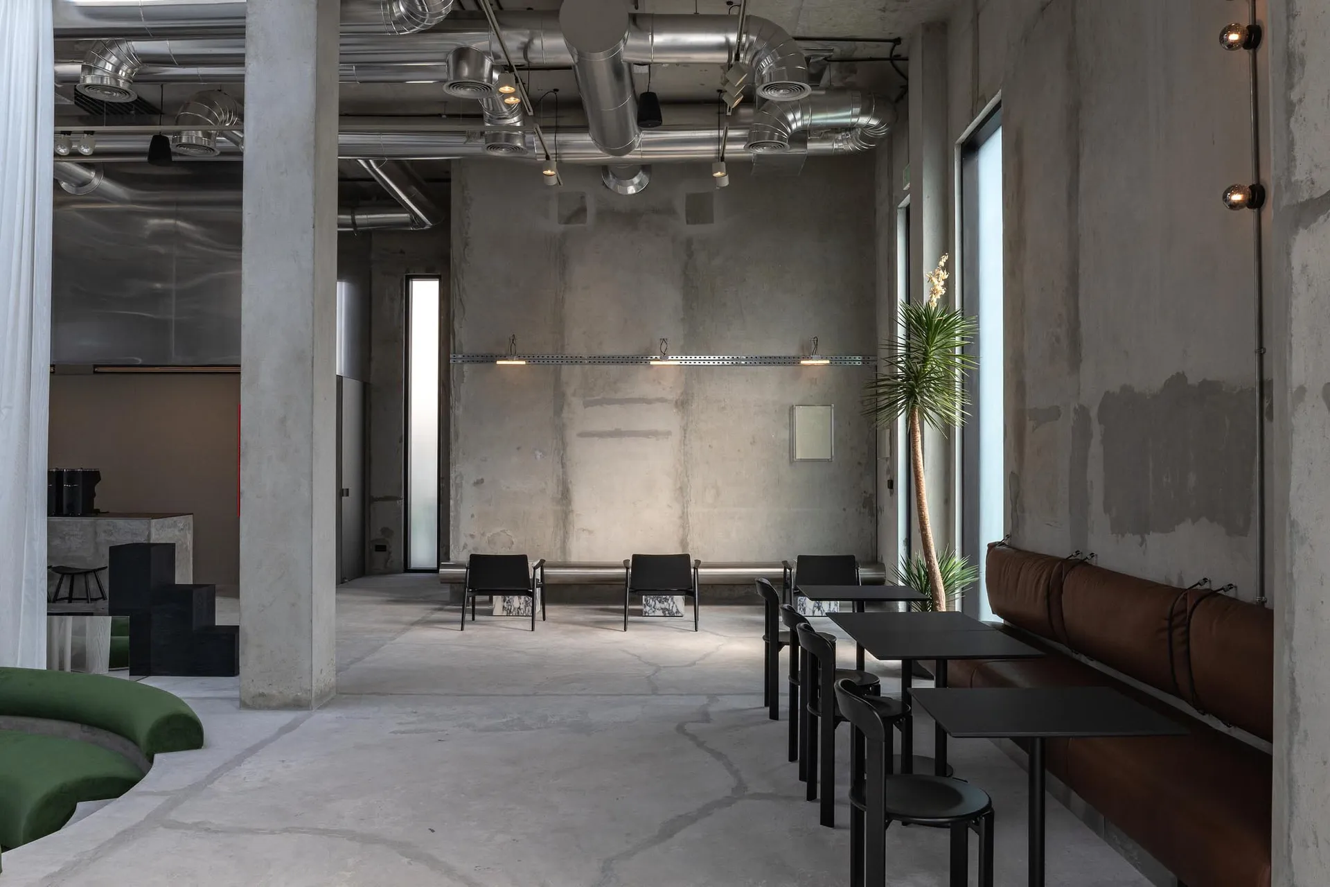寬敞的工業風咖啡廳室內空間,展現清水模牆面、銀色通風管線、棕色皮革長椅、黑色餐桌,以及前景的弧形綠色模組沙發。