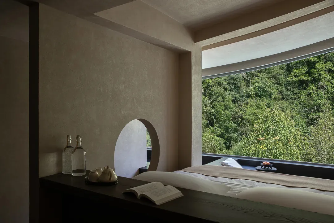 Modern minimalist bedroom interior with textured plaster walls, dark wood console, open book, and large window overlooking a green mountain forest.