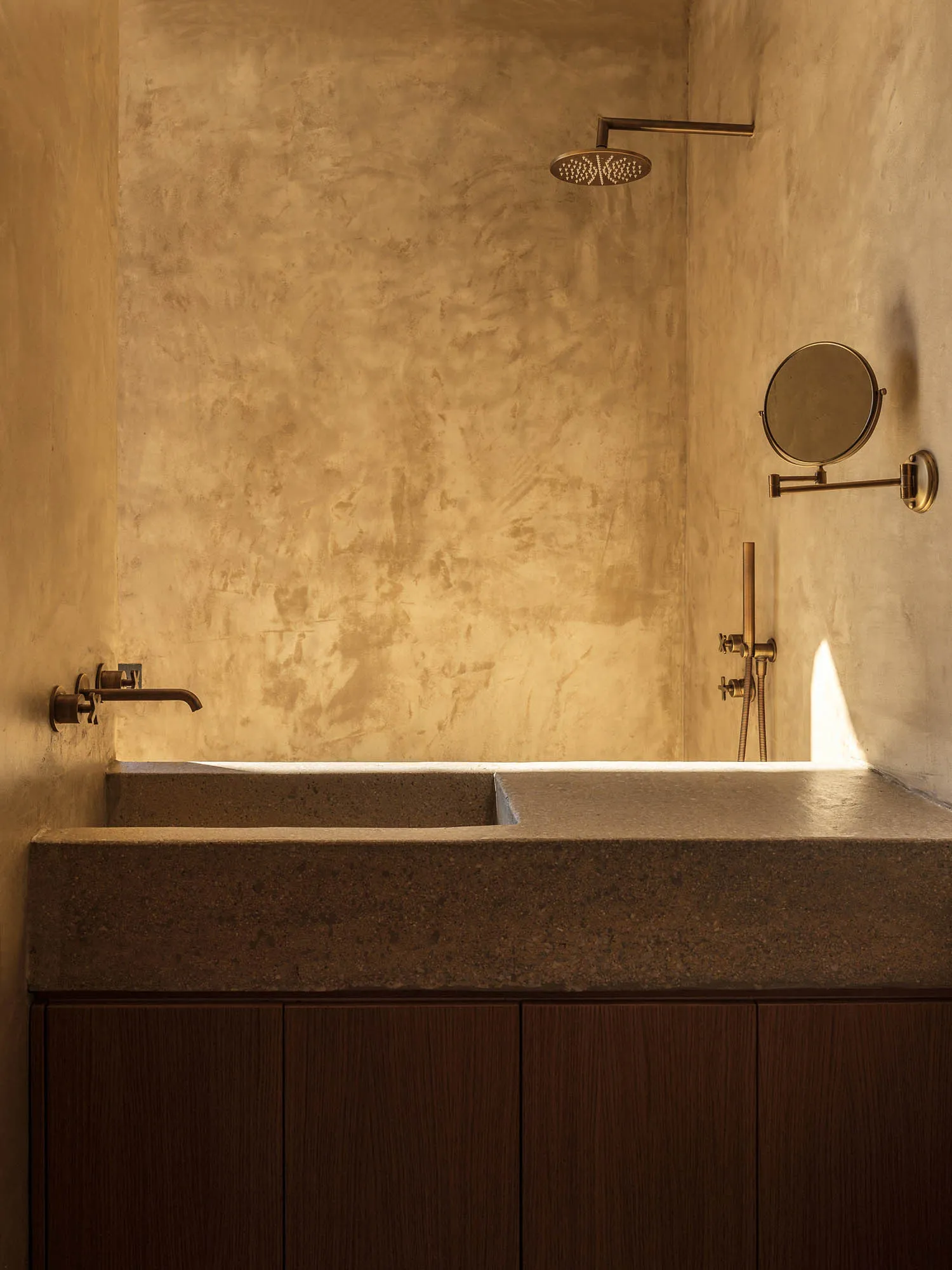 Modern minimalist bathroom featuring a concrete shower-bath, textured plaster walls, and warm brass fixtures with a round magnifying mirror.