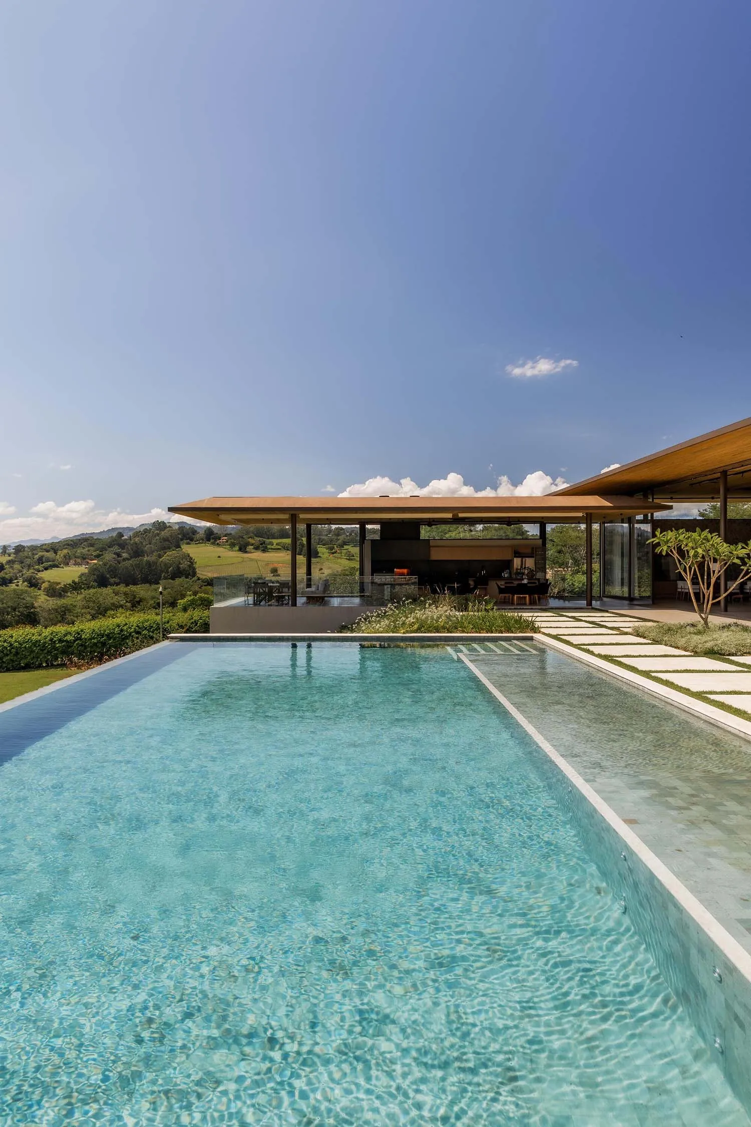 Turquoise infinity pool with travertine deck extending toward a modern open-plan villa with large glass walls, set against a scenic rural landscape under a bright blue sky.