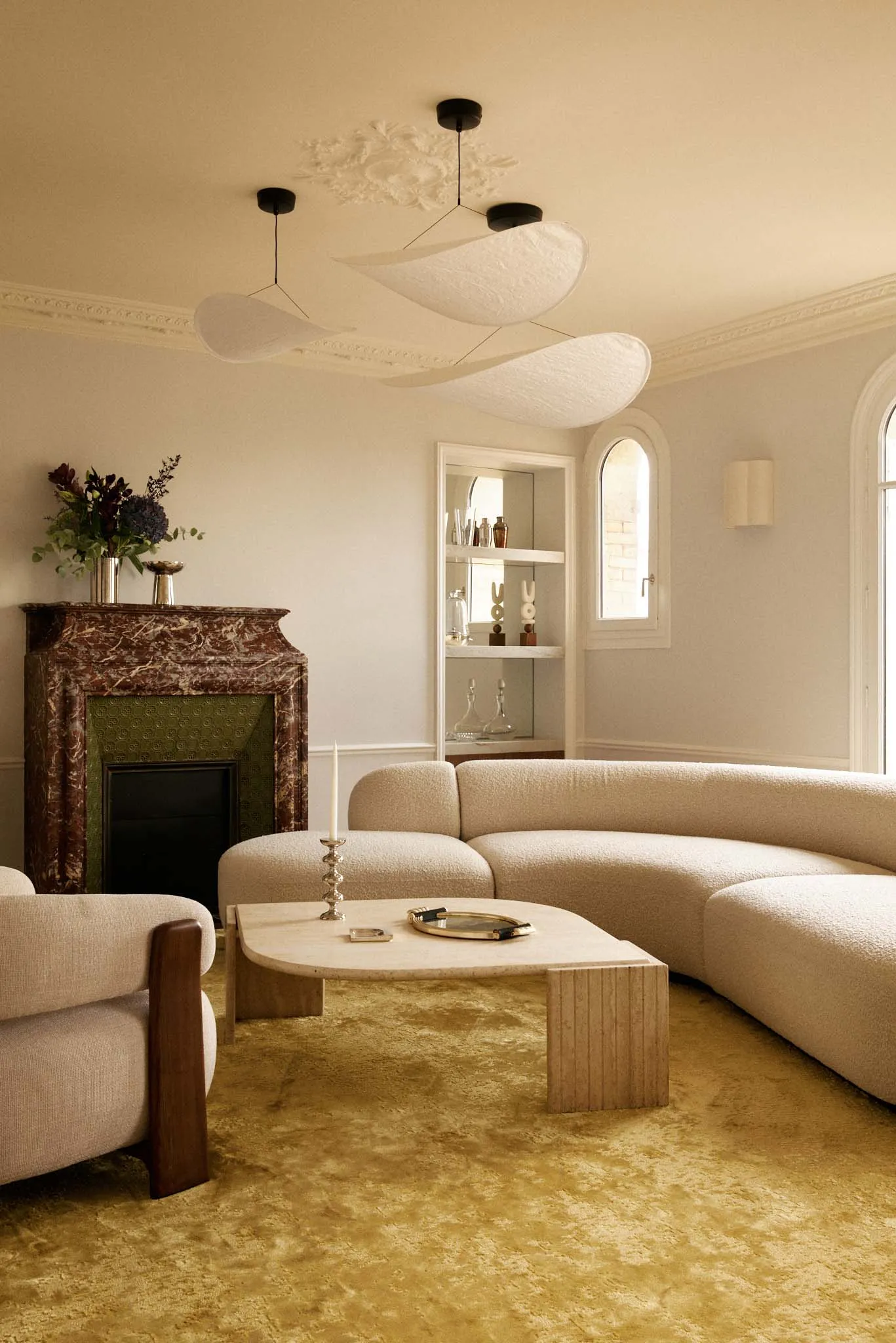 Elegant living room featuring a large curved bouclé sofa, marble fireplace with green tile interior, travertine coffee table, and yellow shaggy carpet under modern sculptural pendant lights.