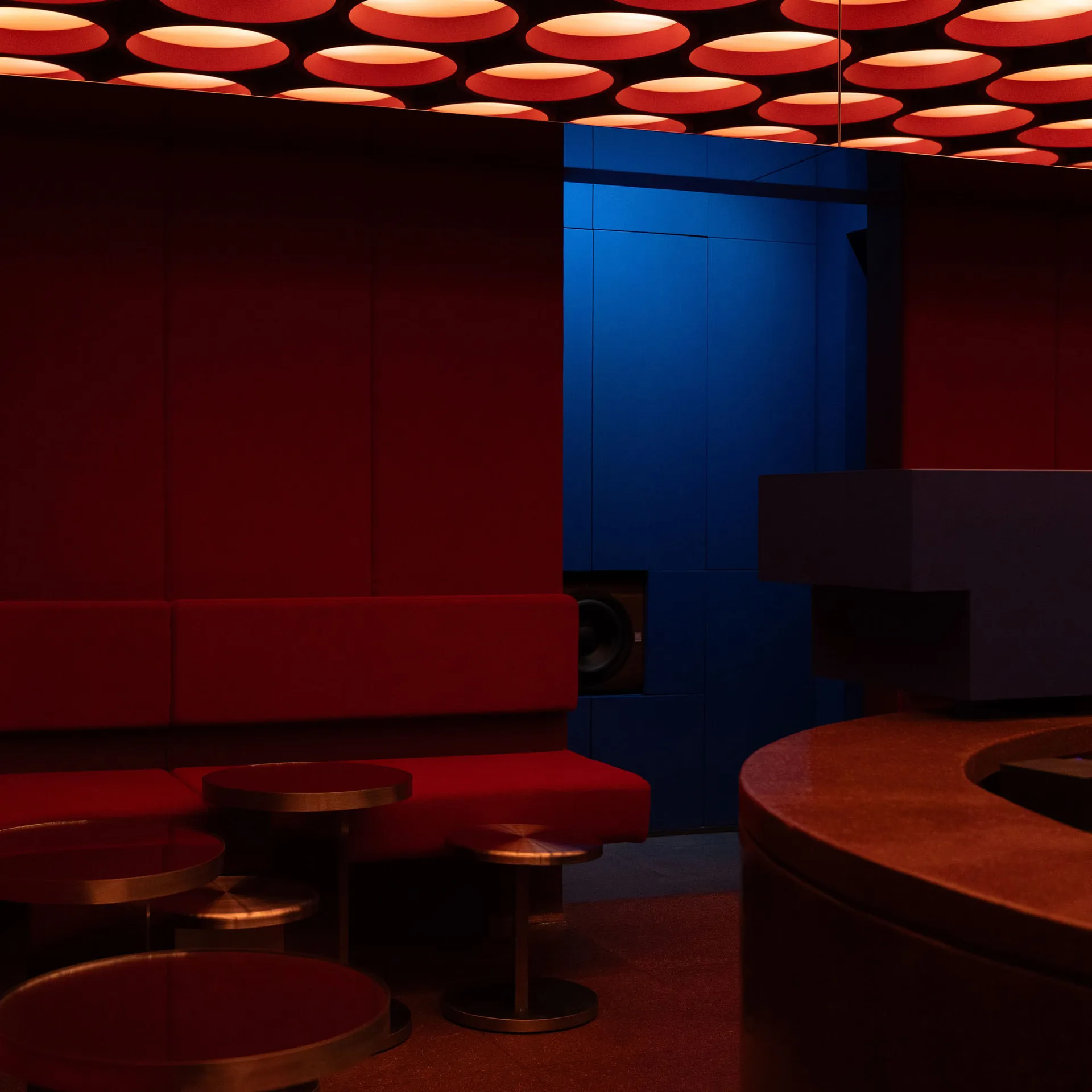 Interior of a modern nightclub lounge with red velvet banquette seating, round metallic tables, and a ceiling covered in glowing red circular light fixtures against a deep blue wall.