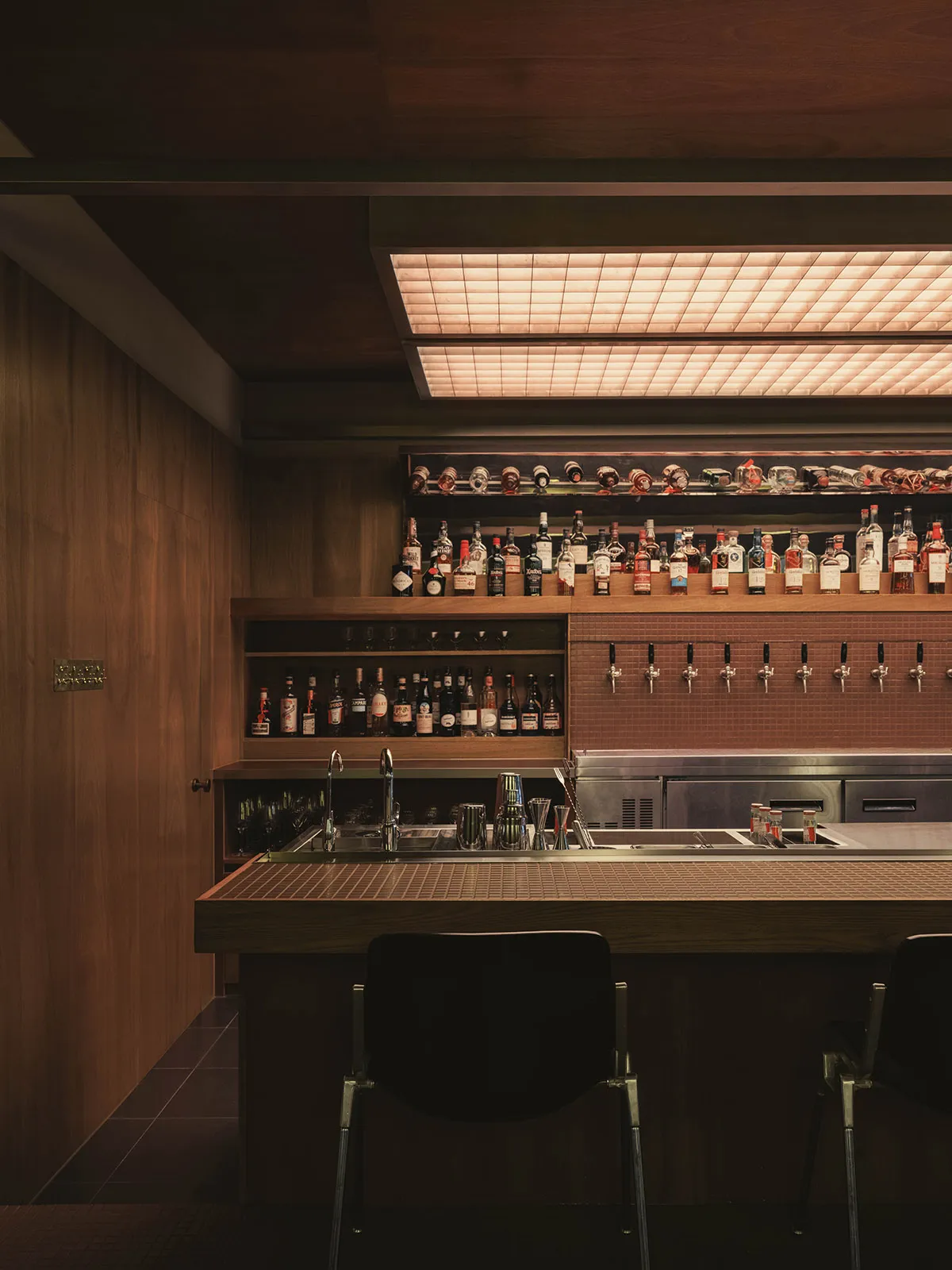 現代家庭酒吧，設有胡桃木護牆板、內嵌式酒瓶展示架、不鏽鋼啤酒龍頭，以及發光格柵天花板燈具。
