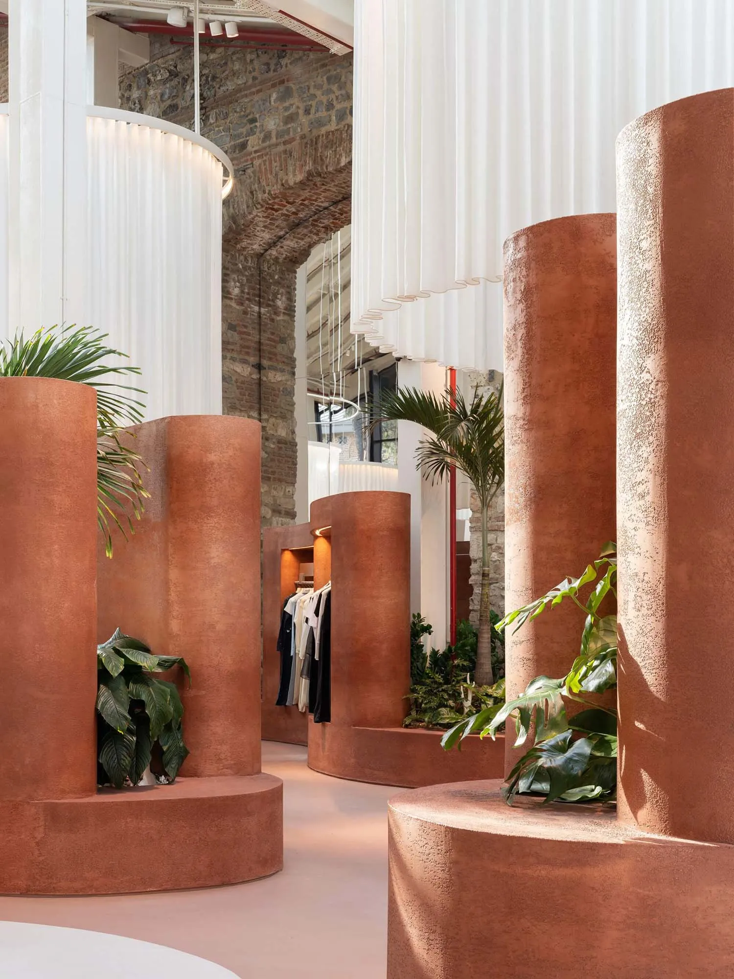 Interior view of a modern boutique featuring large curved terracotta plaster columns, tropical houseplants, white sheer curtains, and exposed brick walls in the background.