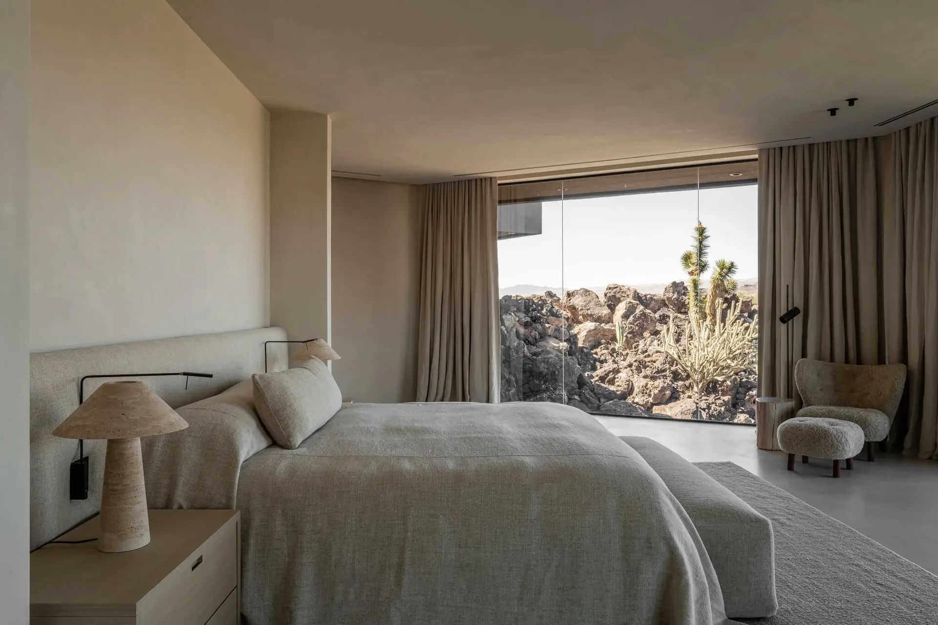 Modern minimalist bedroom with neutral linen bedding, travertine table lamp, and large glass windows overlooking a rocky desert landscape with cacti.