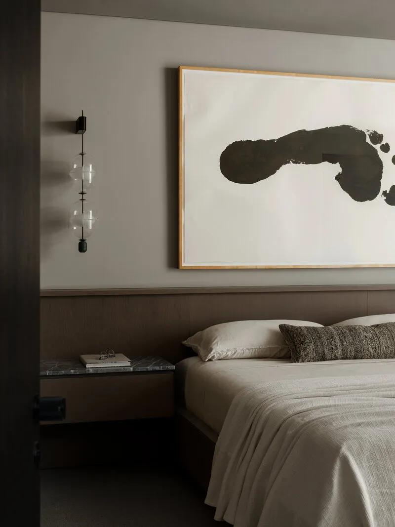 Modern minimalist bedroom featuring a walnut wood panelled headboard, neutral linen bedding, marble nightstand, modern glass wall sconce, and large abstract black ink painting.