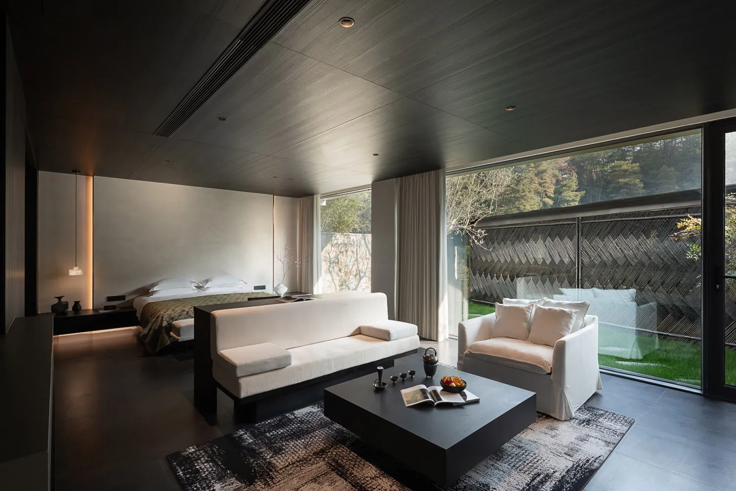 Modern minimalist bedroom sitting area featuring a white sofa, white armchair, dark wood coffee table, and large floor-to-ceiling windows overlooking a private garden with a patterned fence.