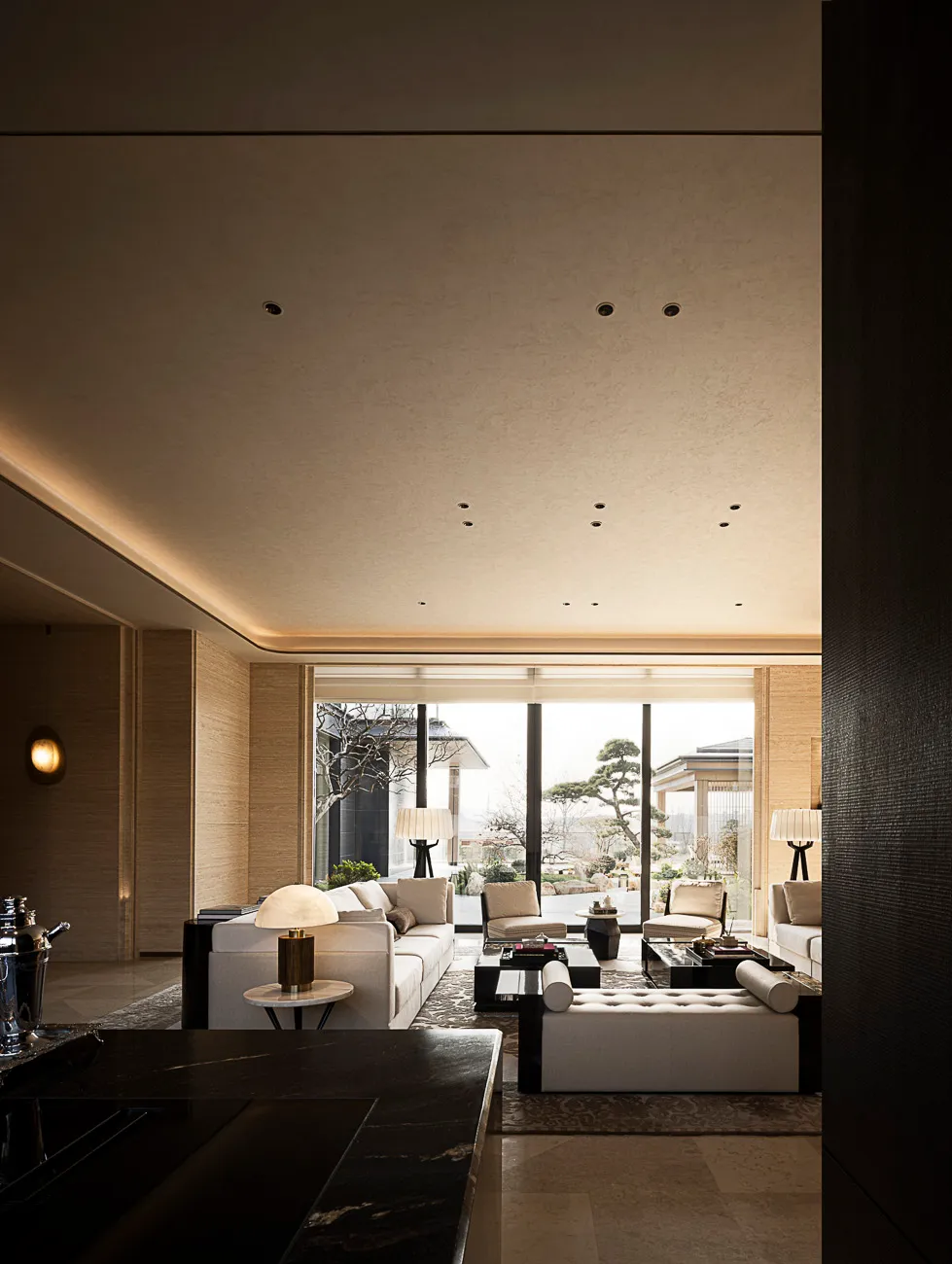 Modern luxury living room featuring travertine walls, large white sectional sofa, dark marble coffee tables, and floor-to-ceiling windows overlooking a Japanese garden with bonsai trees.
