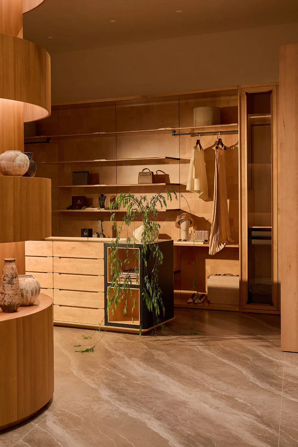Luxurious walk-in closet featuring custom light oak cabinetry, a curved wooden display island, hanging plants, marble-look tiled floor, and soft warm lighting.