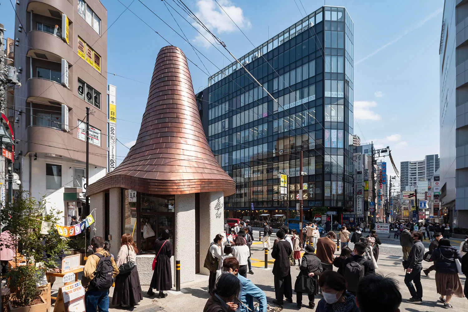 Daytime view of a distinctive copper conical roof building surrounded by pedestrians on a busy street in Kichijoji, Tokyo, with modern glass buildings in the background.