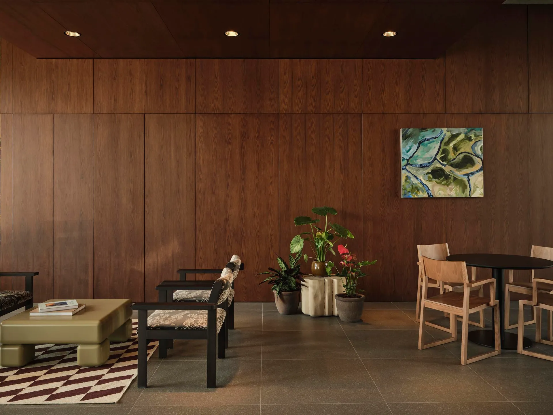 Modern lounge area with dark walnut wood panel walls, black-framed patterned armchairs, olive leather ottoman on a geometric rug, round black dining table with light wood chairs, and multiple potted plants under recessed ceiling lights.