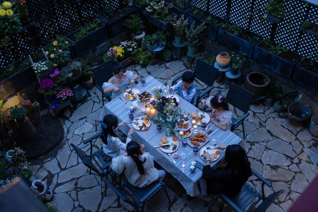 Overhead view of a family of six enjoying dinner at a long table set on a rustic stone-paved courtyard at dusk, surrounded by numerous potted flowers and plants.