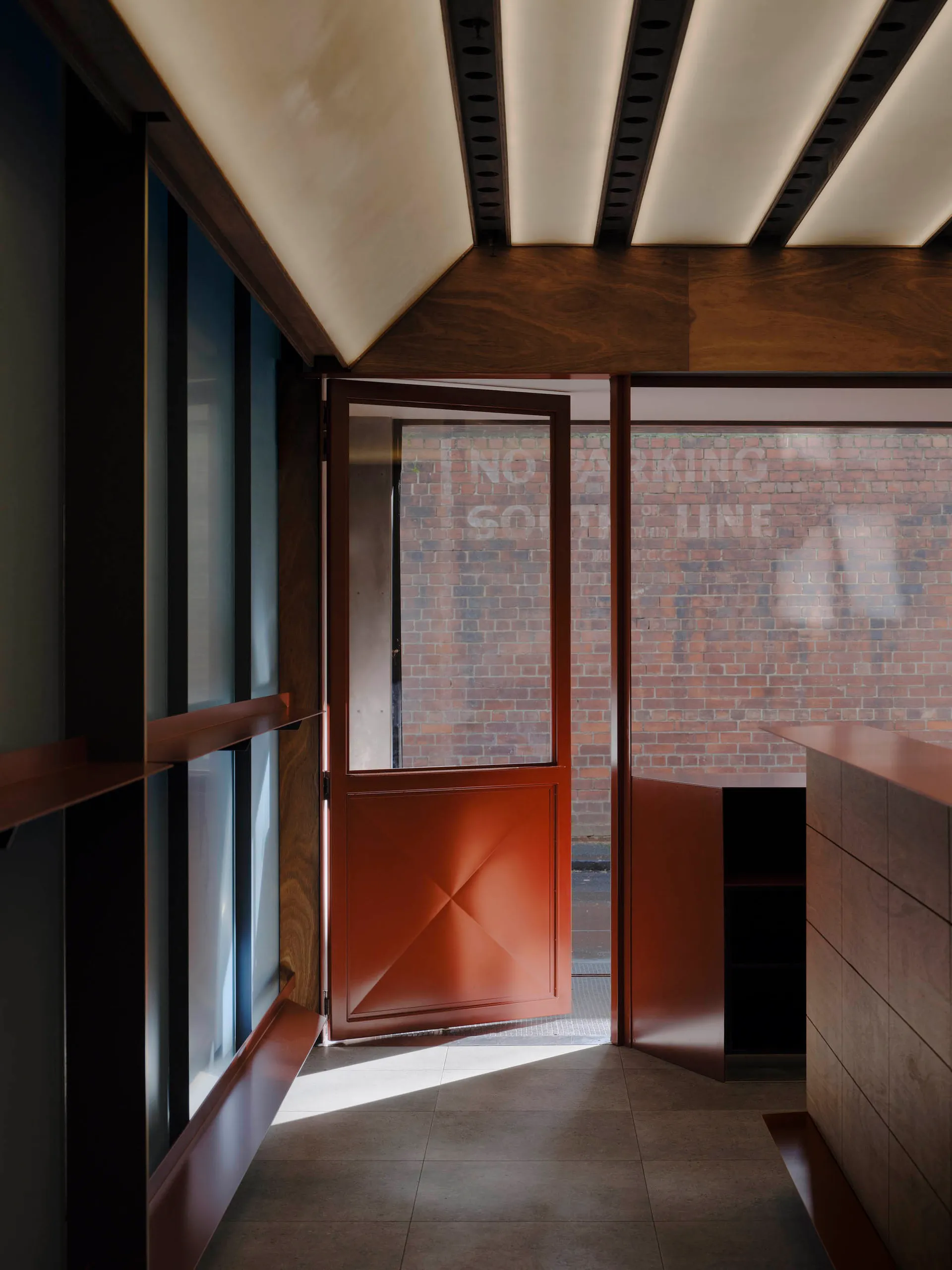 Modern minimalist entryway featuring dark walnut wood paneling on walls and ceiling, a terracotta-colored geometric pivot door standing open, and a view of a brick wall outside.