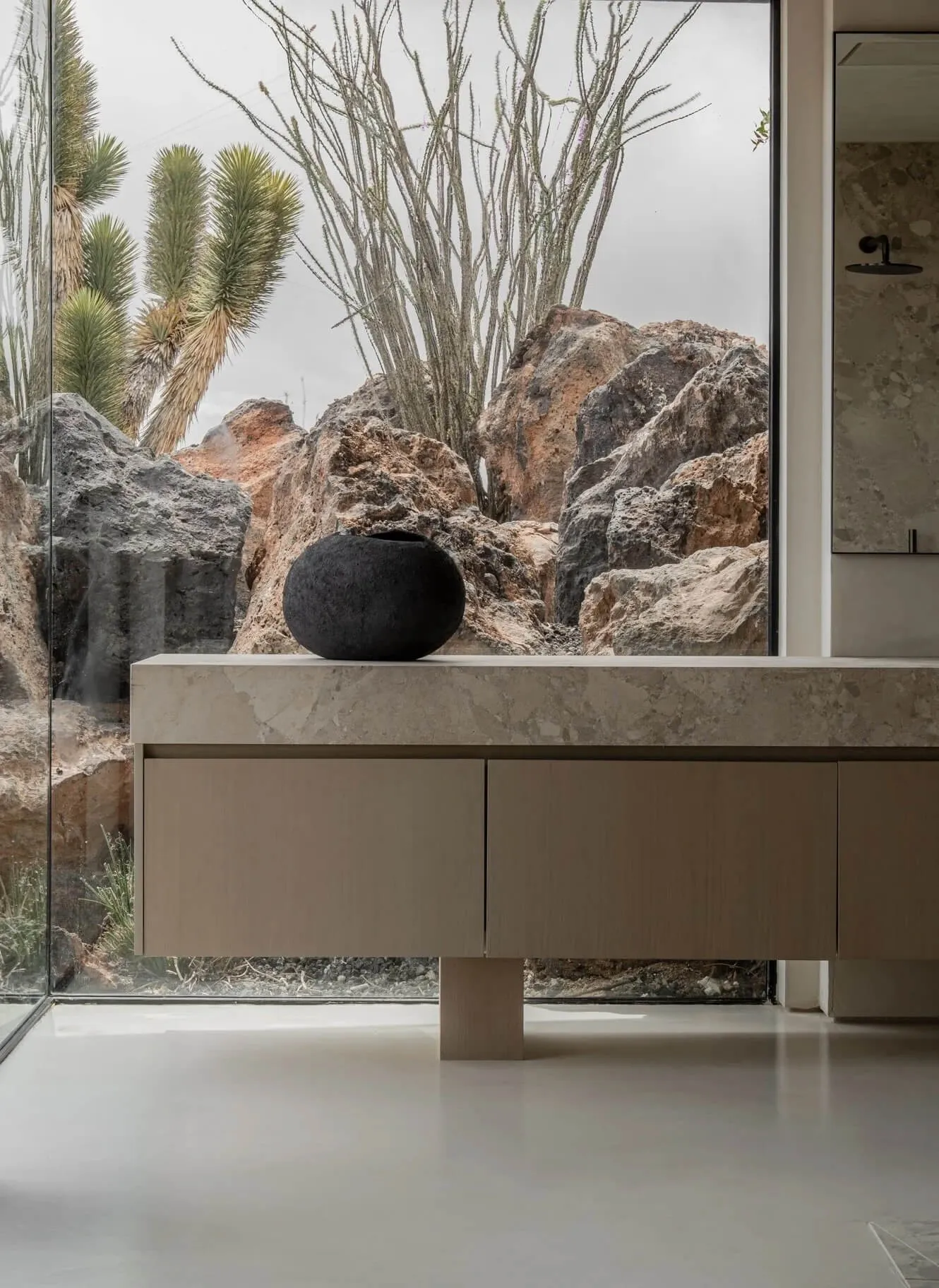 Modern minimalist bathroom featuring a floating travertine vanity with a black ceramic vessel, large glass window overlooking dramatic desert rocks and cacti.
