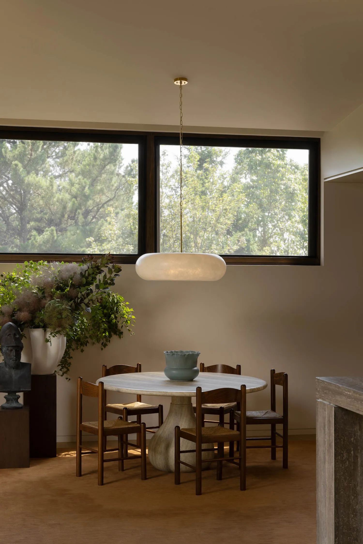 Modern dining room featuring a round travertine table, four dark wood woven-seat chairs, a large alabaster pendant lamp, and a large window overlooking green trees.