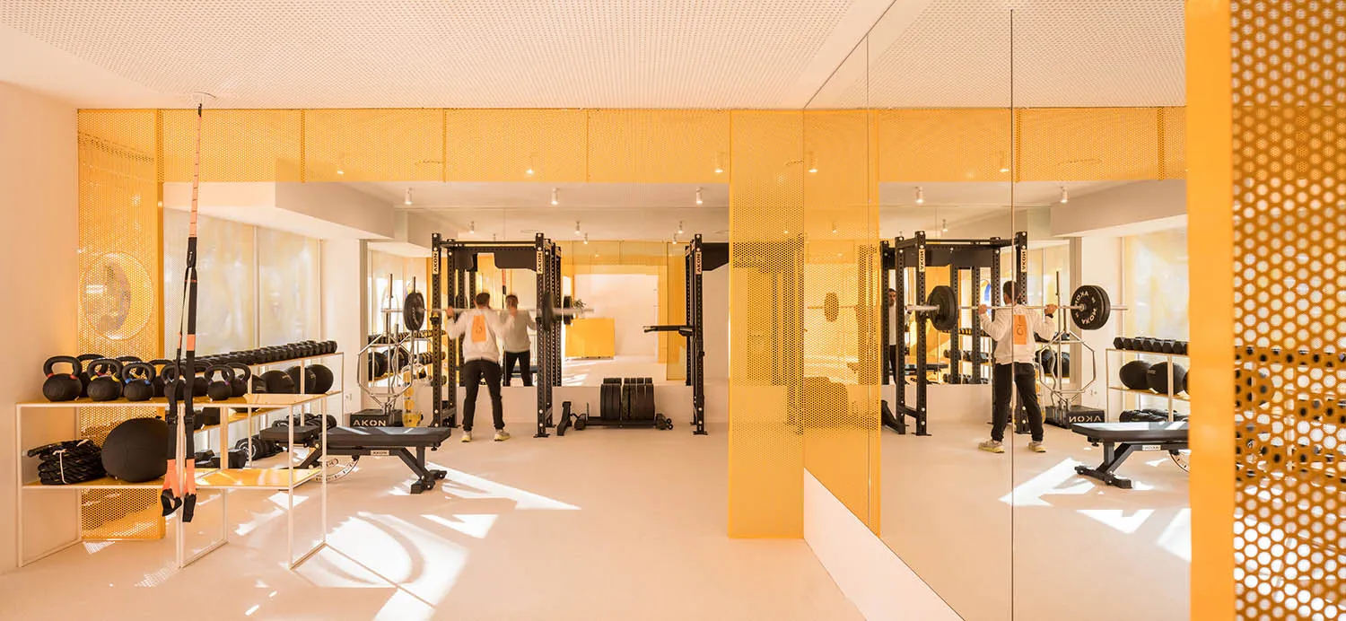 Modern home gym interior featuring bright yellow perforated metal room dividers, large mirrors, weight racks, benches, and two people training with barbells.