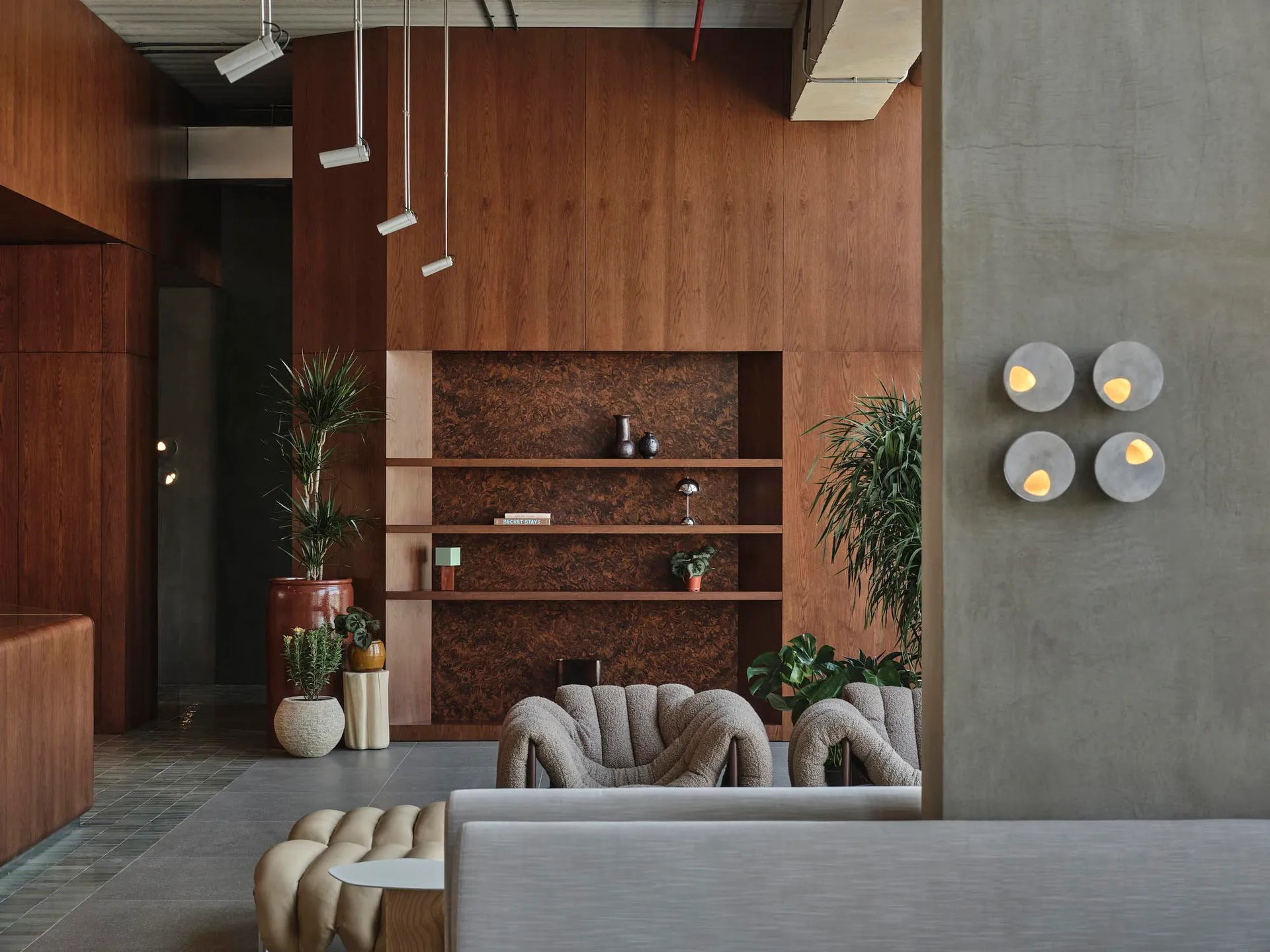 Modern lobby interior featuring a large walnut burl wood feature wall, built-in shelving, two curved bouclé armchairs, concrete wall sconces, and multiple potted plants.