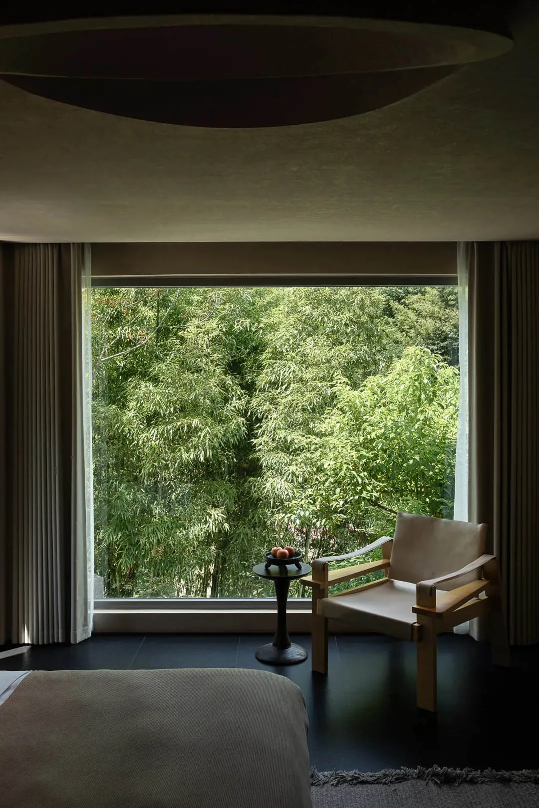 Modern bedroom interior featuring a wooden and linen armchair next to a large window overlooking a lush green bamboo forest, with dark flooring and curved ceiling details.