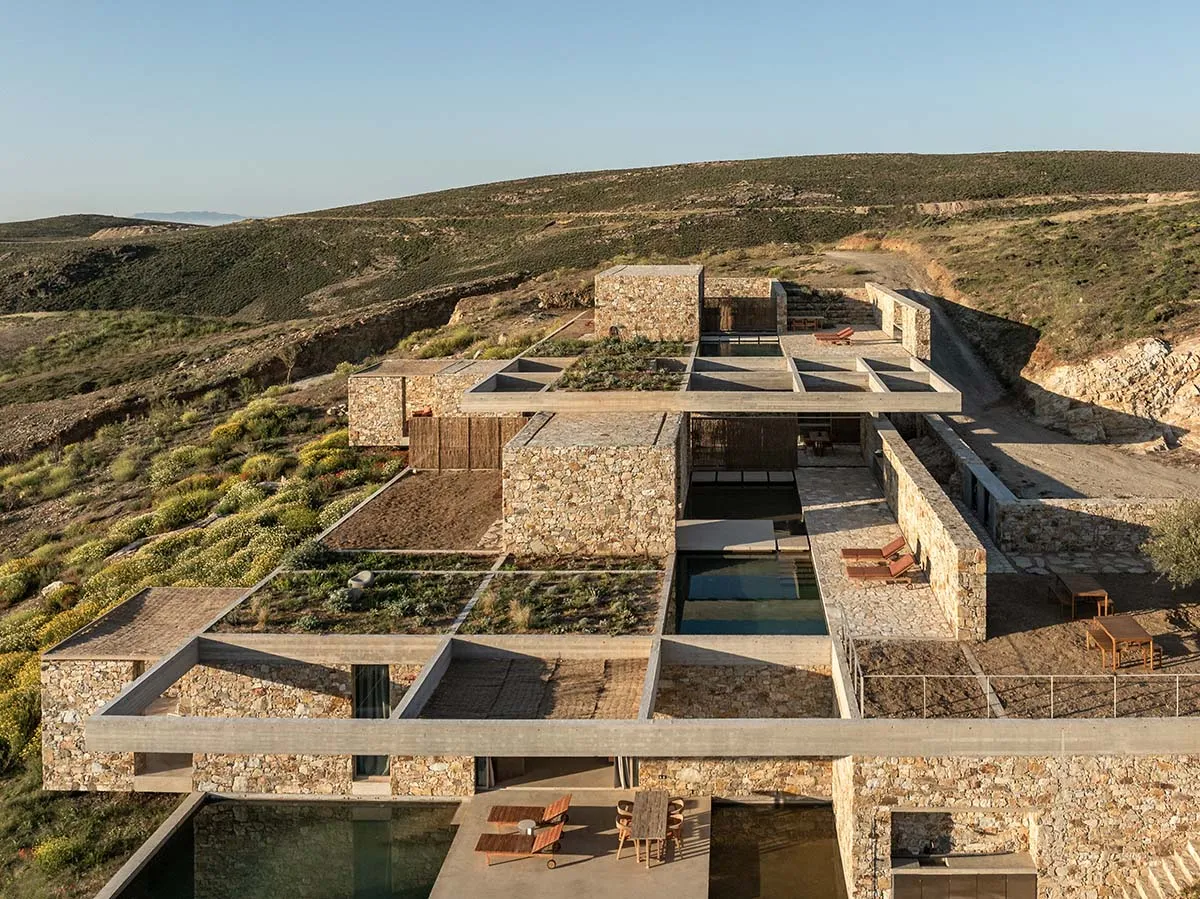 Aerial view of a contemporary stone and concrete courtyard villa with multiple terraces, infinity pools, and green roofs integrated into a rugged Mediterranean hillside.
