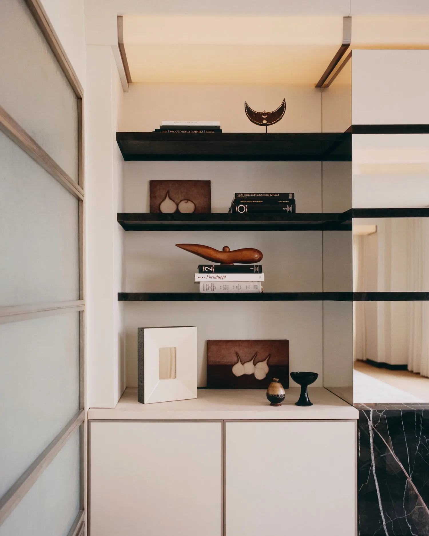 Modern minimalist built-in shelving unit with dark walnut floating shelves, white cabinetry, and carefully arranged books, wooden sculptures, and ceramic objects against a neutral wall.