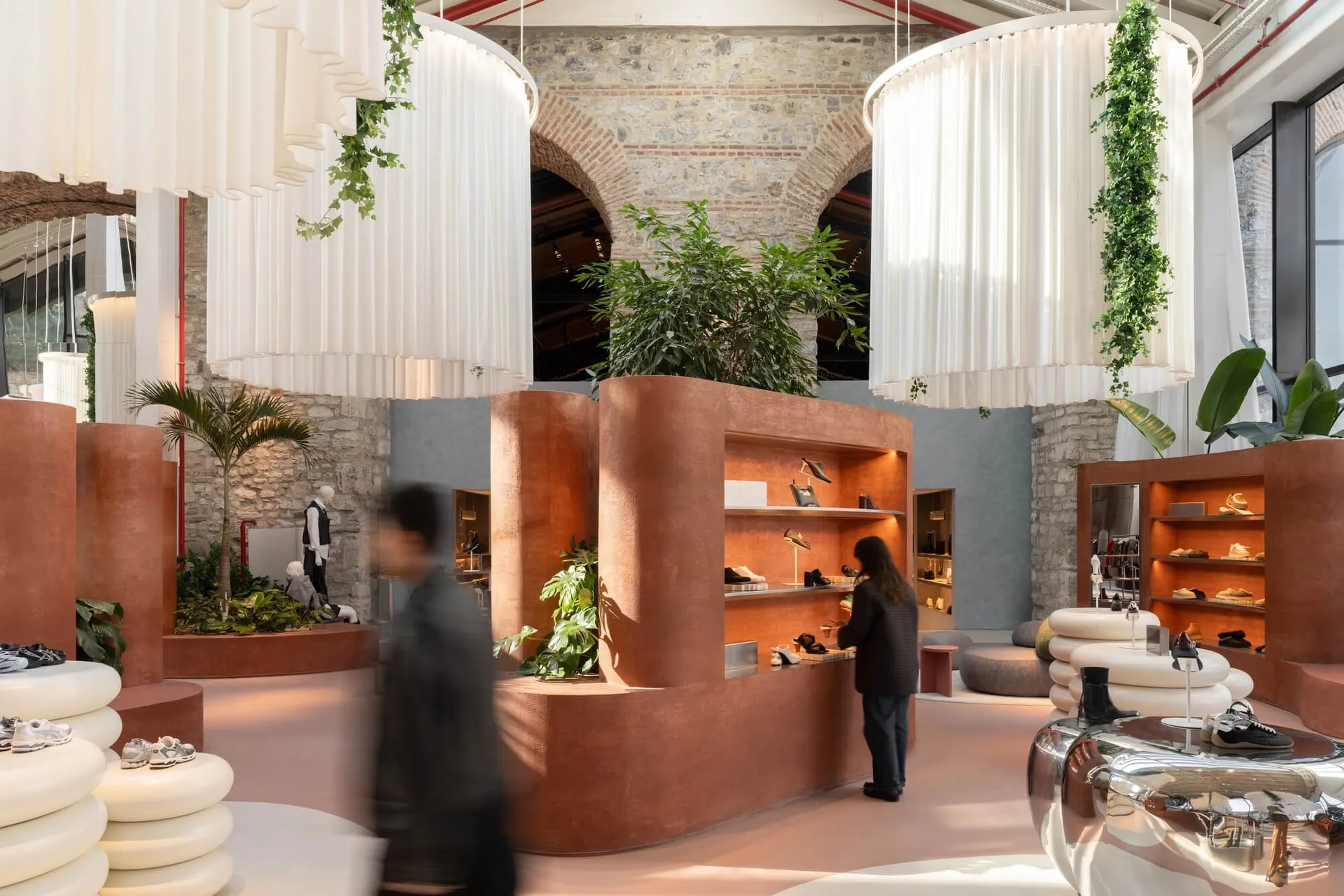 Modern shoe store interior featuring curved terracotta display walls, large white cylindrical fabric pendant lights with hanging greenery, exposed brick arches, and shoppers browsing sneakers.