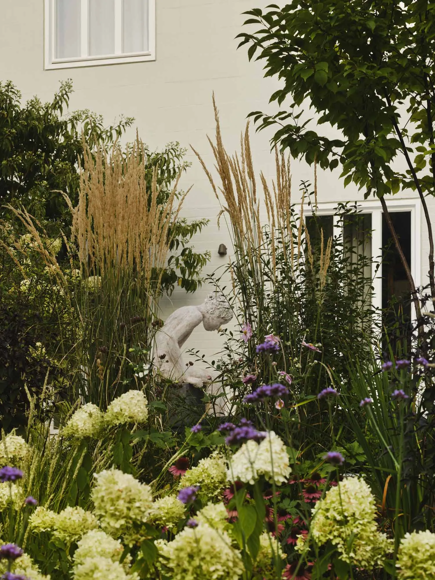 A classical stone sculpture of a kneeling female figure surrounded by tall ornamental grasses, white hydrangeas, purple verbena, and pink coneflowers in front of a white house wall.