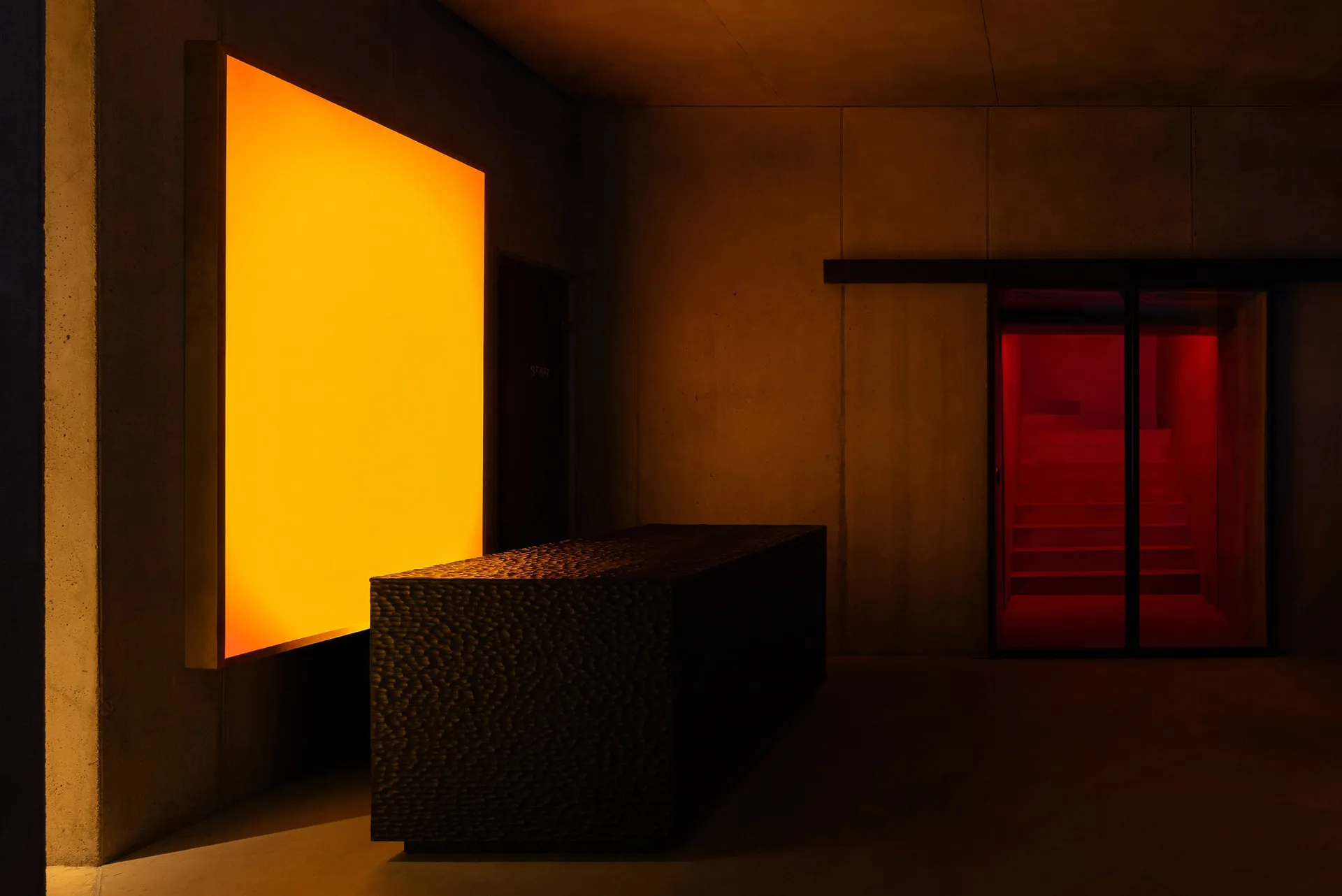Brutalist concrete interior featuring a textured concrete bench, a glowing warm orange light panel on the left wall, and a red illuminated staircase visible through glass doors on the right.