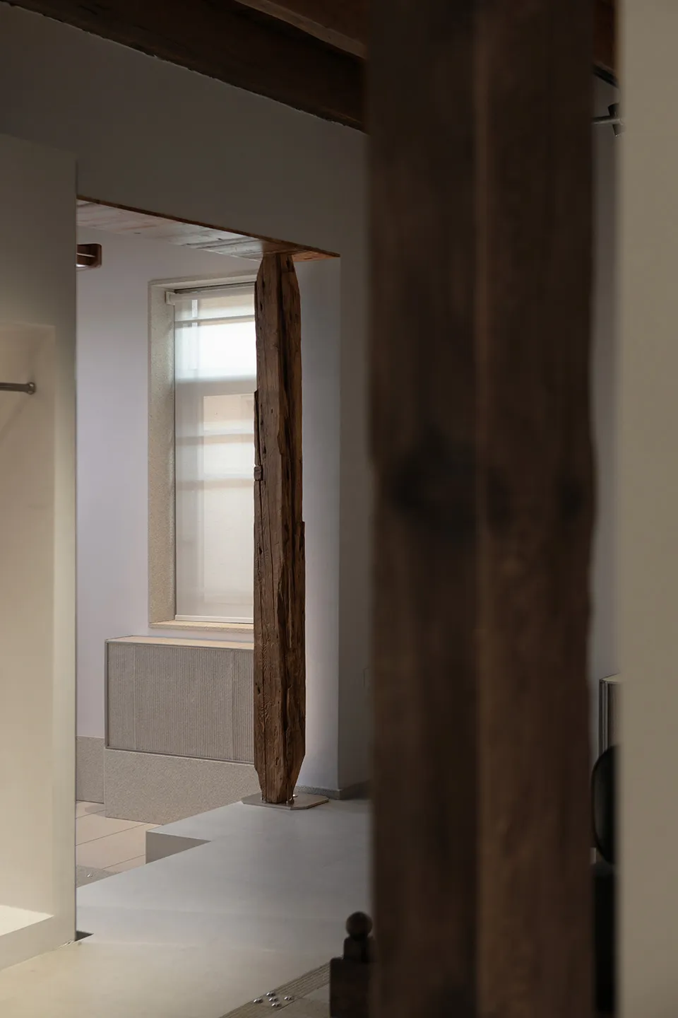 Vertical reclaimed wood sculpture standing in a bright, minimalist interior hallway with white walls and concrete floor.
