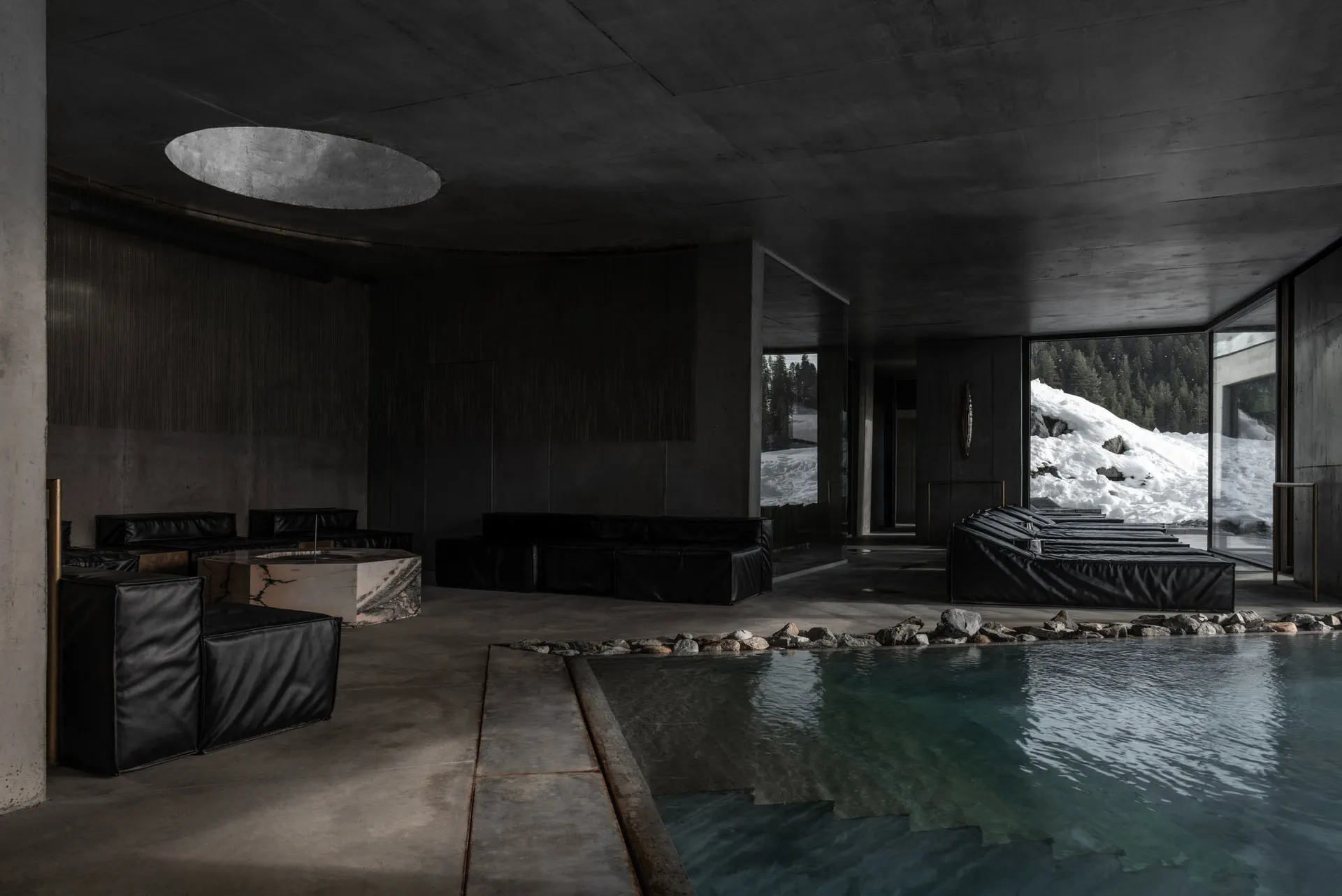 Modern brutalist indoor swimming pool with black leather modular sofas, marble tables, and large glass windows revealing a snowy mountain forest outside.