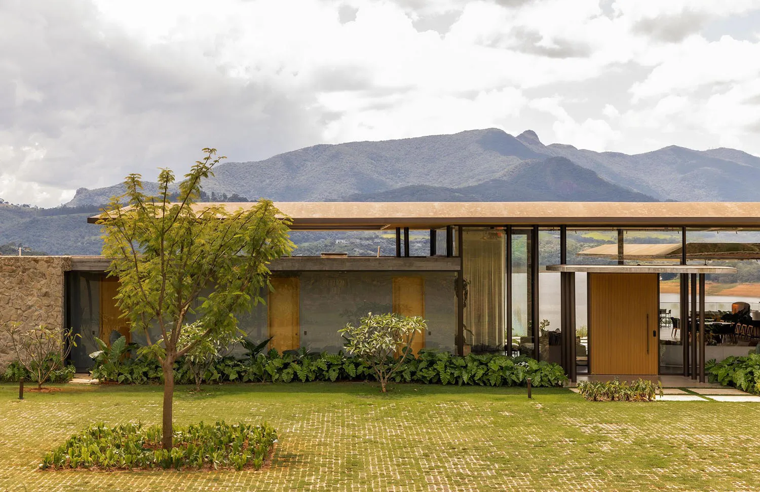 Modern single-story mountain house featuring concrete walls, vertical wood slats, large glass windows, and lush tropical landscaping with a dramatic mountain range in the background.