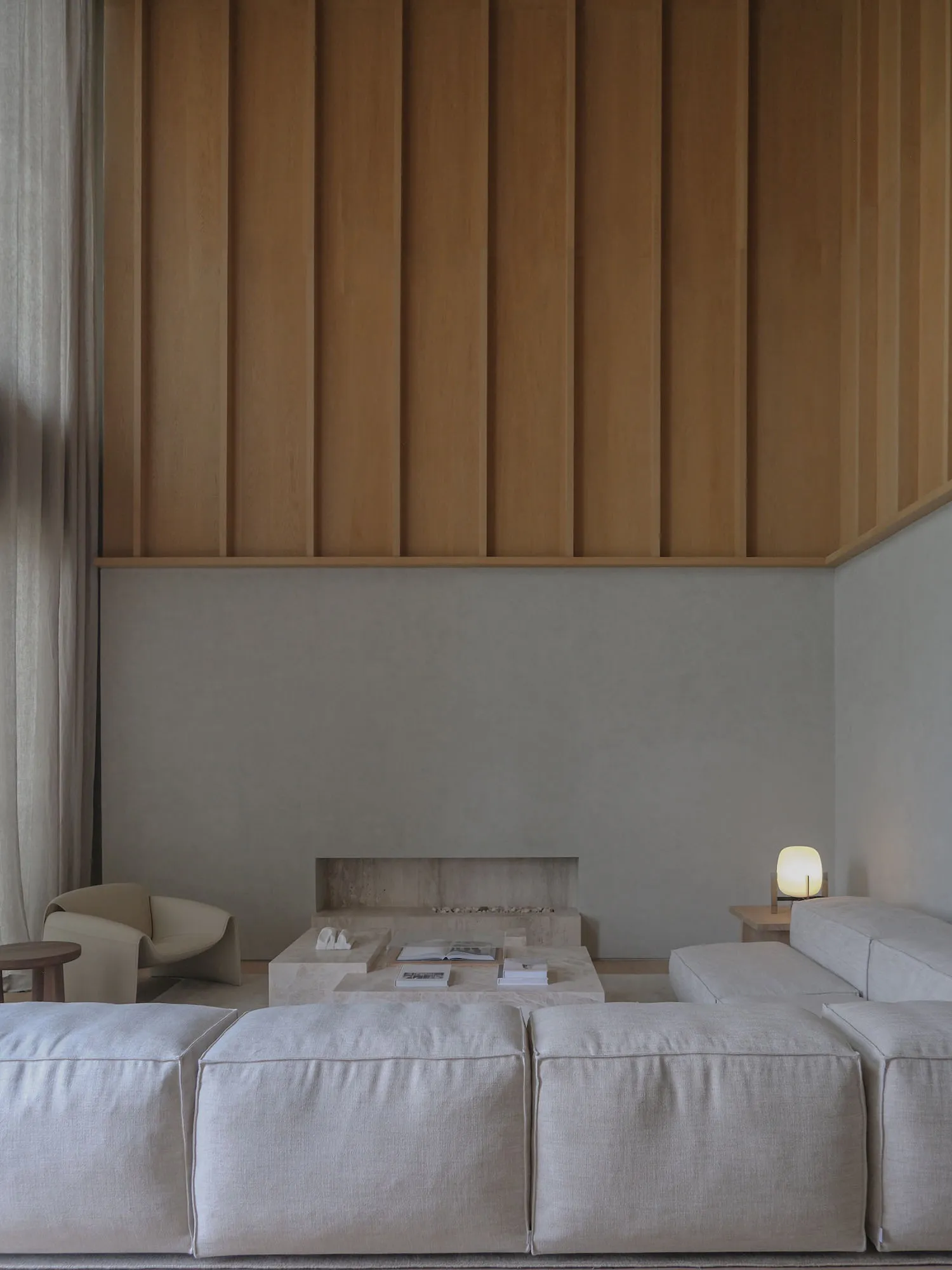 Modern minimalist living room featuring a large modular beige sofa, travertine coffee tables, a built-in stone hearth, and a tall oak wood slatted accent wall under soft natural light.