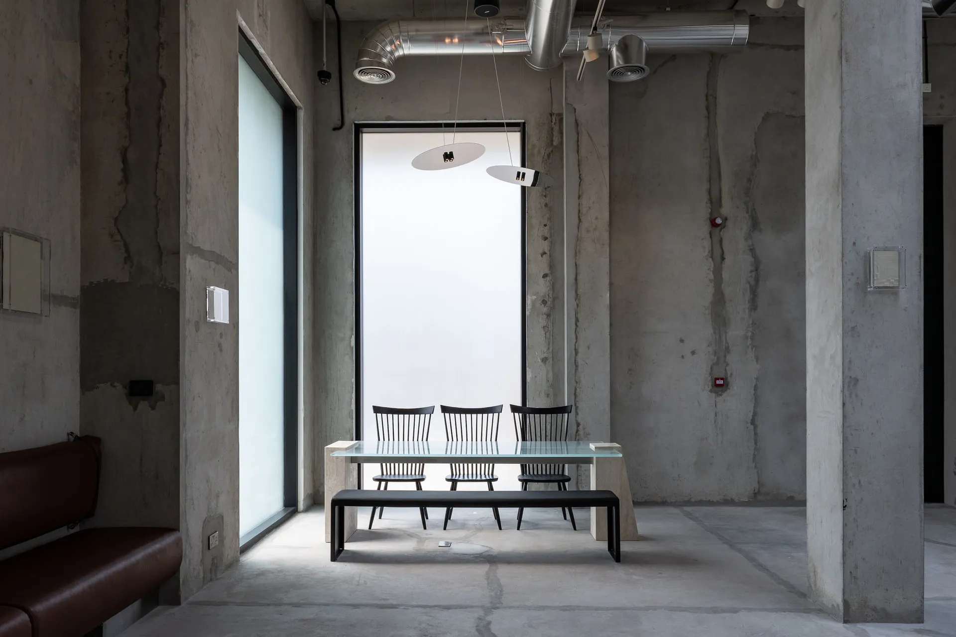 Industrial-style dining area featuring raw concrete walls, a glass dining table with black spindle-back chairs, a black bench, and two modern pendant lights suspended from an exposed ceiling with visible ductwork.